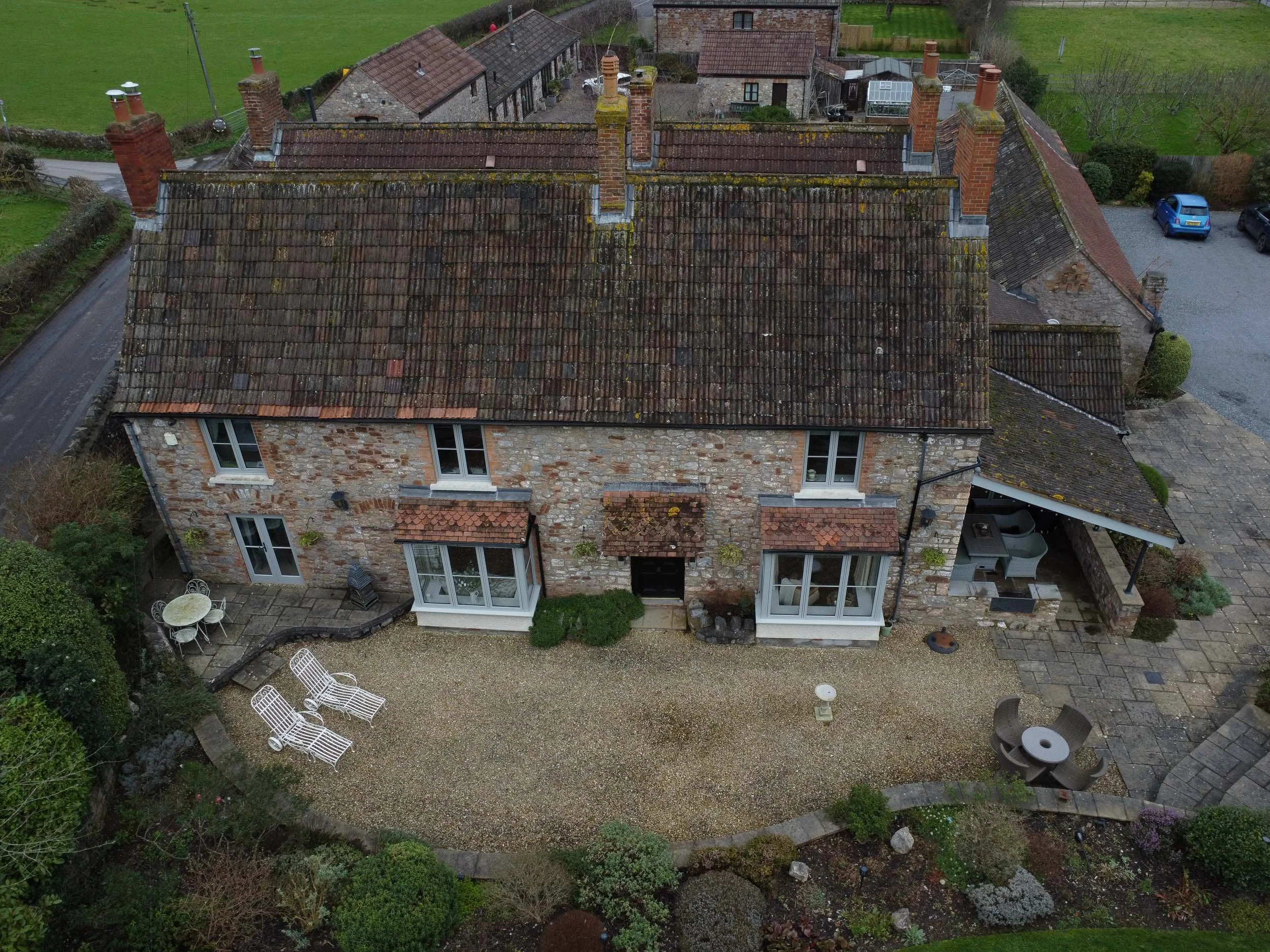 Aerial view of a brick house with a gravel backyard and outdoor furniture, surrounded by garden and neighboring buildings.