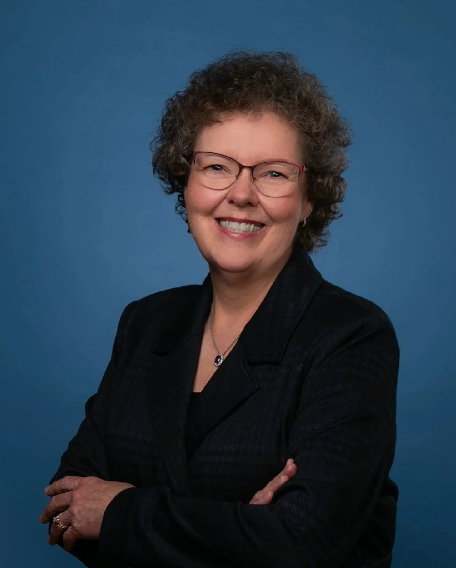 A smiling middle-aged woman with curly hair, wearing glasses, a black blazer, and a pendant necklace, standing against a blue background.