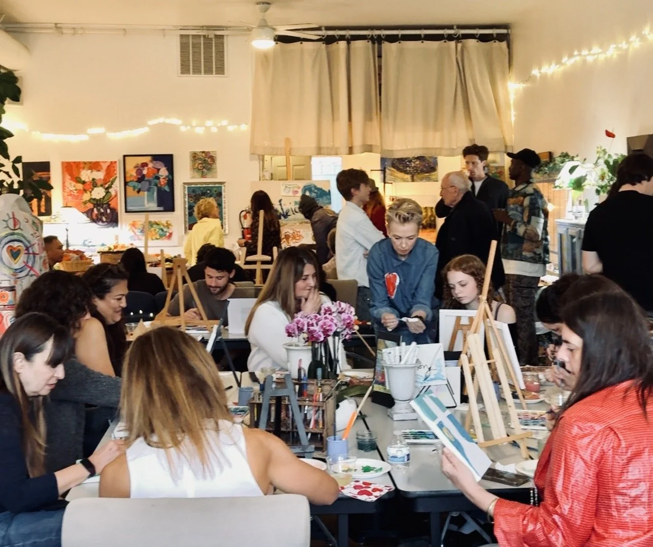 People participating in a group painting event inside a decorated art studio with paintings on the walls, string lights, and various art supplies on tables.