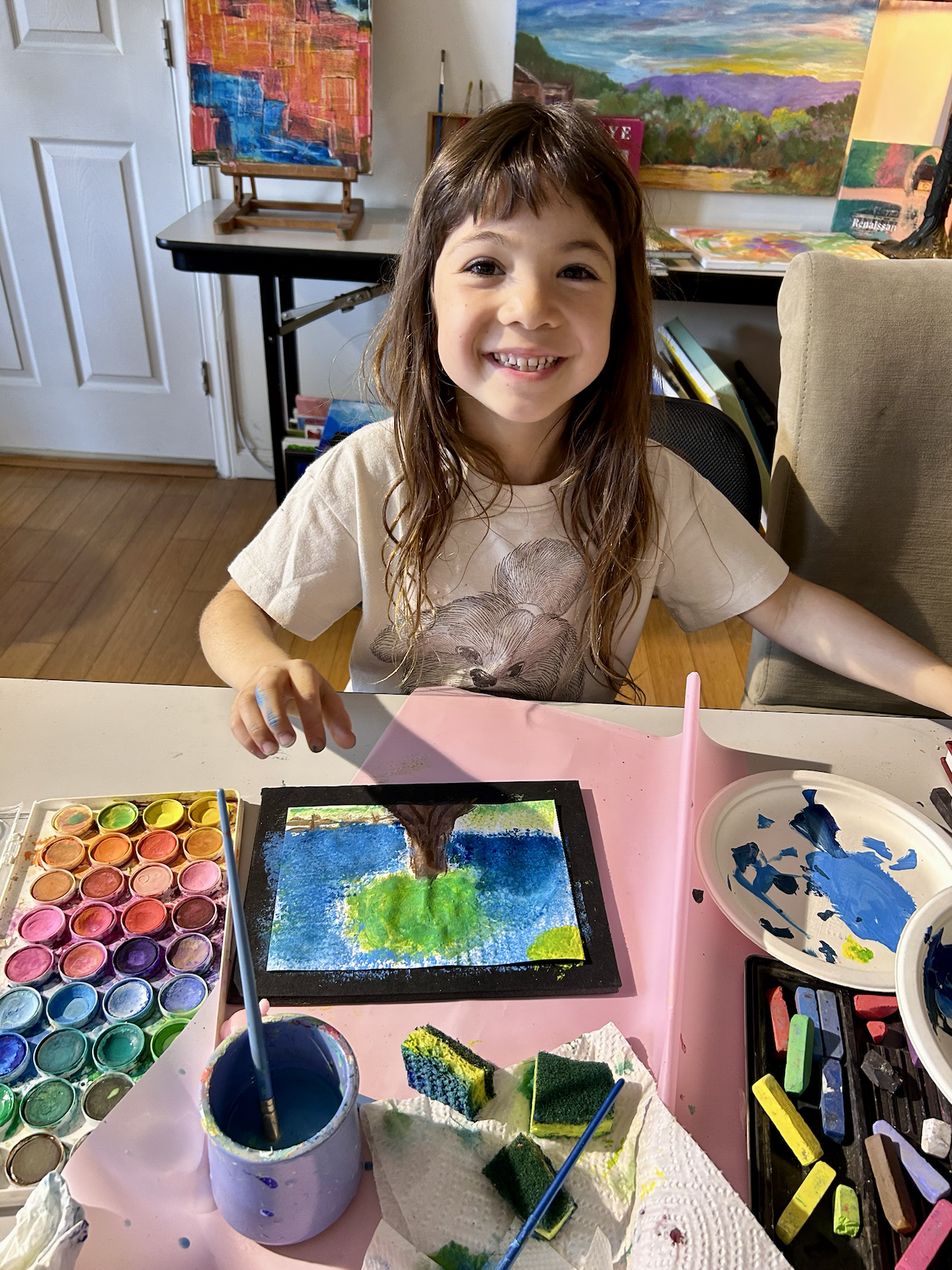A young girl smiles at the camera while sitting at a table with watercolor painting supplies. She is wearing a beige t-shirt with a dog illustration, and her art features a large tree with green grass and a blue sky. The background includes several colorful landscape paintings and art supplies.