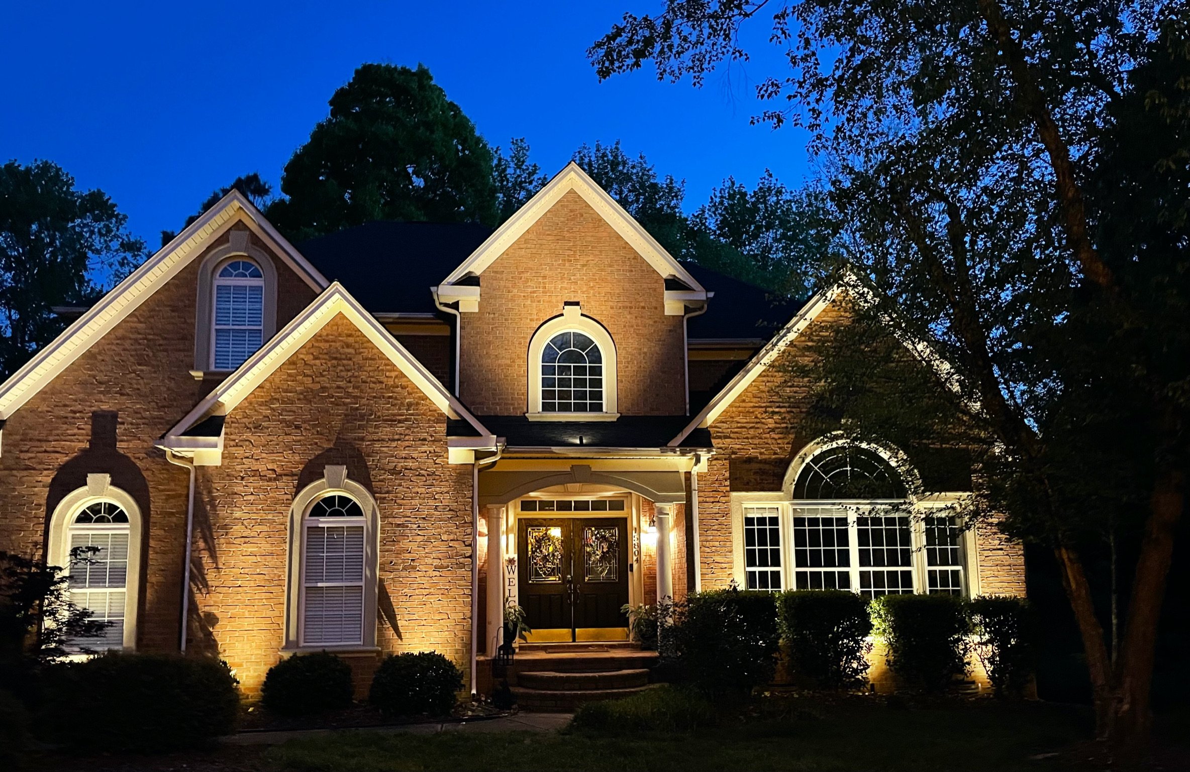 Lake Norman home with custom front elevation landscape lighting, illuminated entryway, warm brick façade uplighting, and professionally designed outdoor lighting after dark.