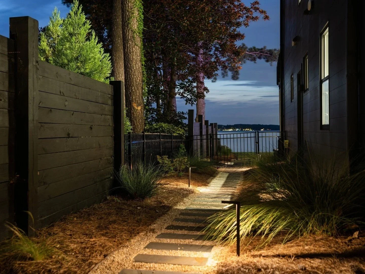 Warm pathway lighting illuminates stepping stones and ornamental grasses along a lakeside garden walkway at dusk.