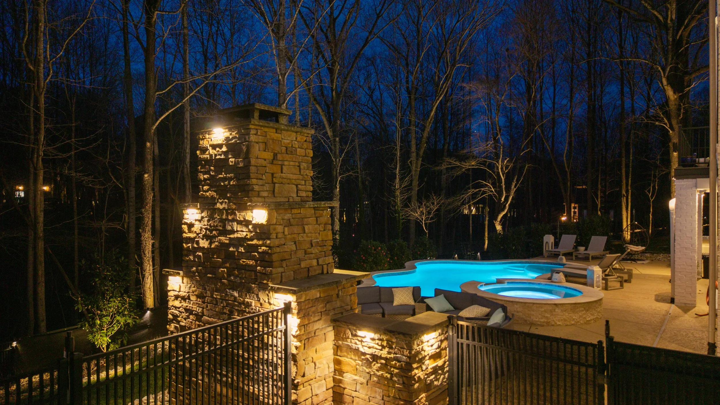 Warm outdoor lighting illuminates a stone fireplace, swimming pool, spa, patio seating, and surrounding woodland at night.