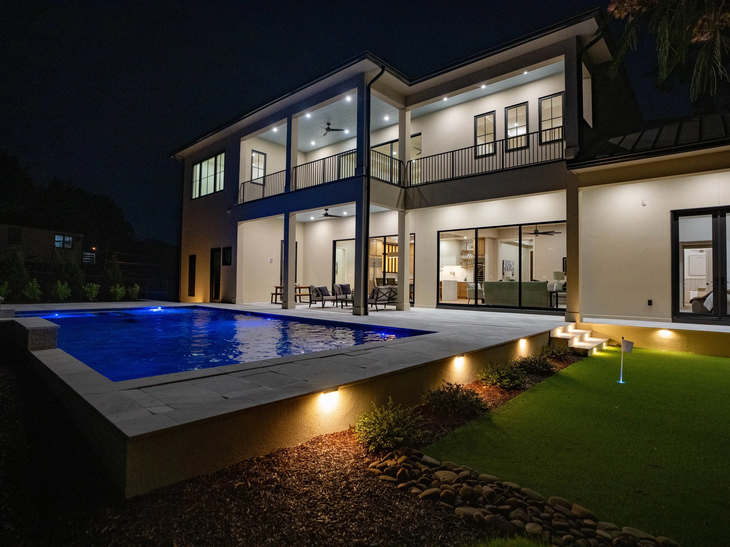 Modern Lake Norman home with a blue-lit pool, covered patios, illuminated steps, retaining walls, and landscape lighting at night.