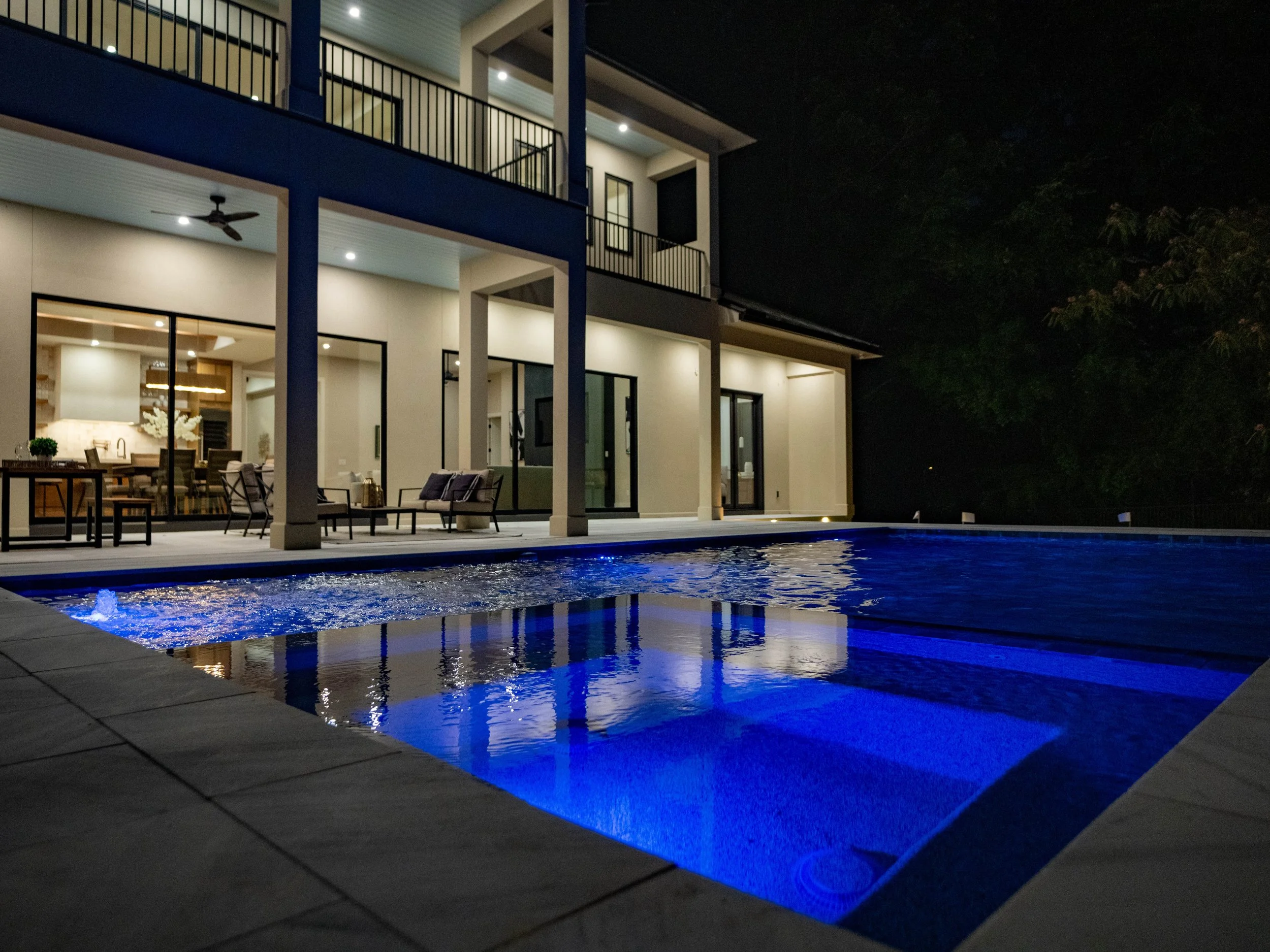 Modern Lake Norman home with a covered patio, illuminated architecture, and a blue-lit swimming pool at night.