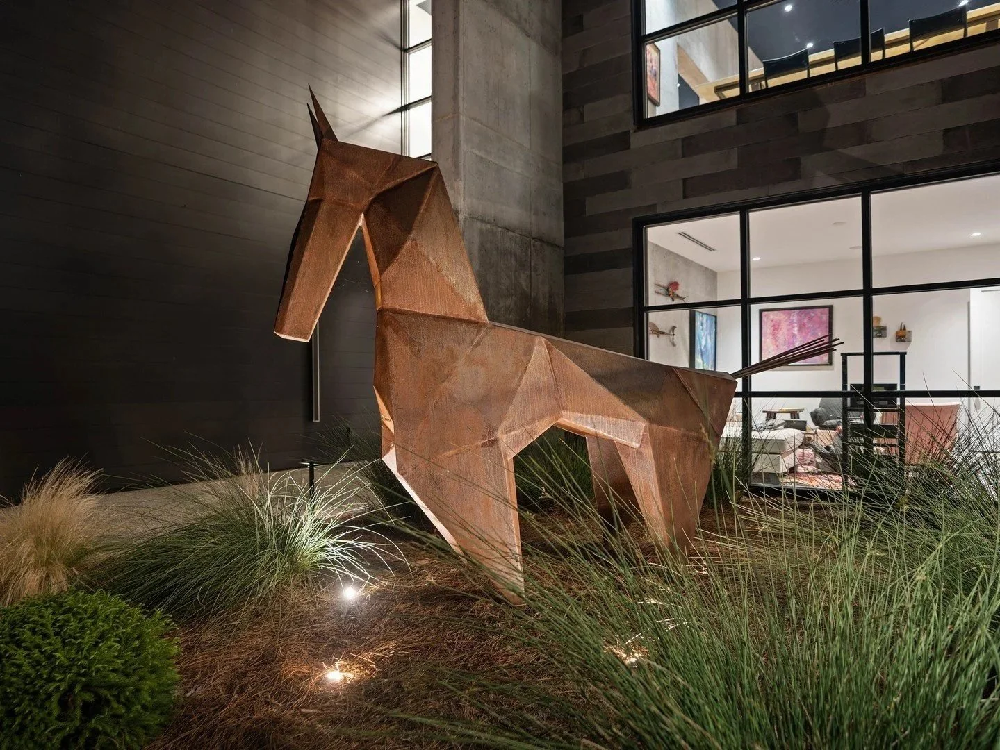 Landscape lighting illuminates a modern metal horse sculpture surrounded by ornamental grasses outside a contemporary home.