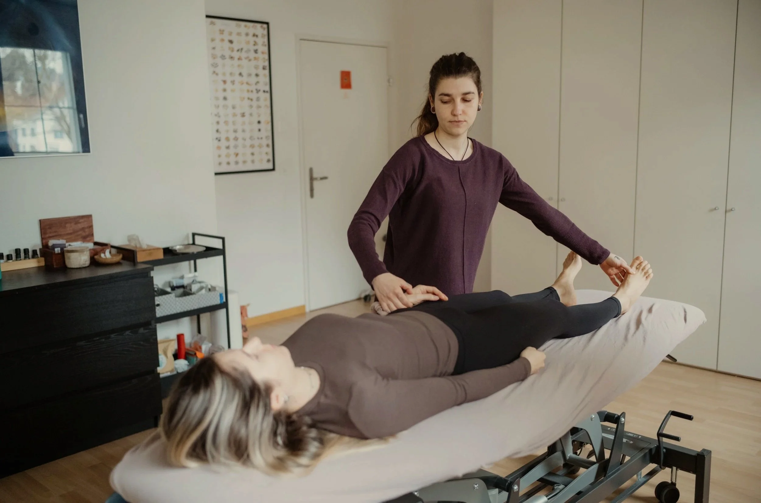 Eine Frau gibt eine Behandlung an den Beinen eines liegenden Patienten in einer Praxis. Im Raum sind Schränke und Poster an der Wand.