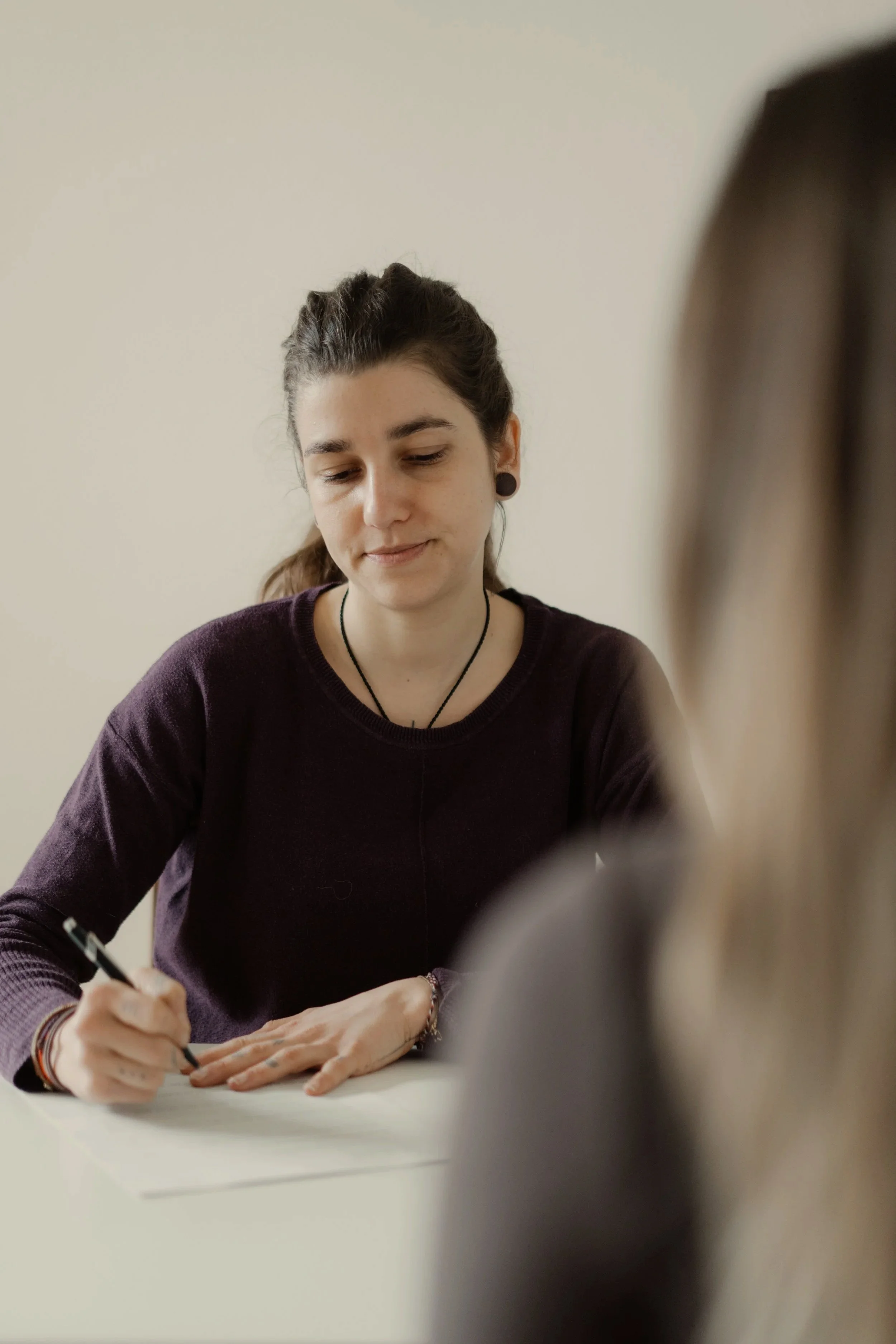 Eine junge lächelnde Frau mit dunklen Haaren in einem Pferdeschwanz sitzt an einem Behandlungstisch in einem hellen Raum und schreibt mit einem schwarzen Stift auf ein Papier.