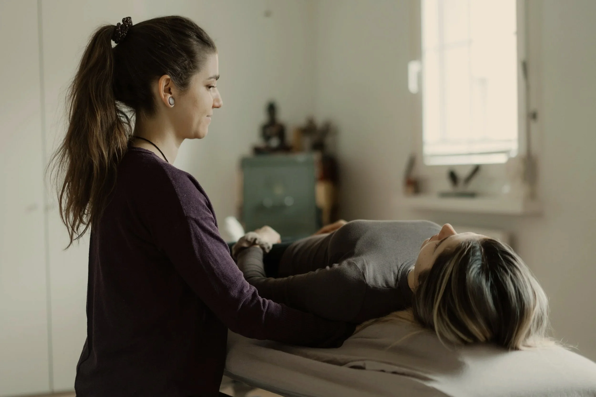 Eine Frau steht neben einer Liege, auf der eine andere Person liegt, während diese therapeutisch behandelt wird.
