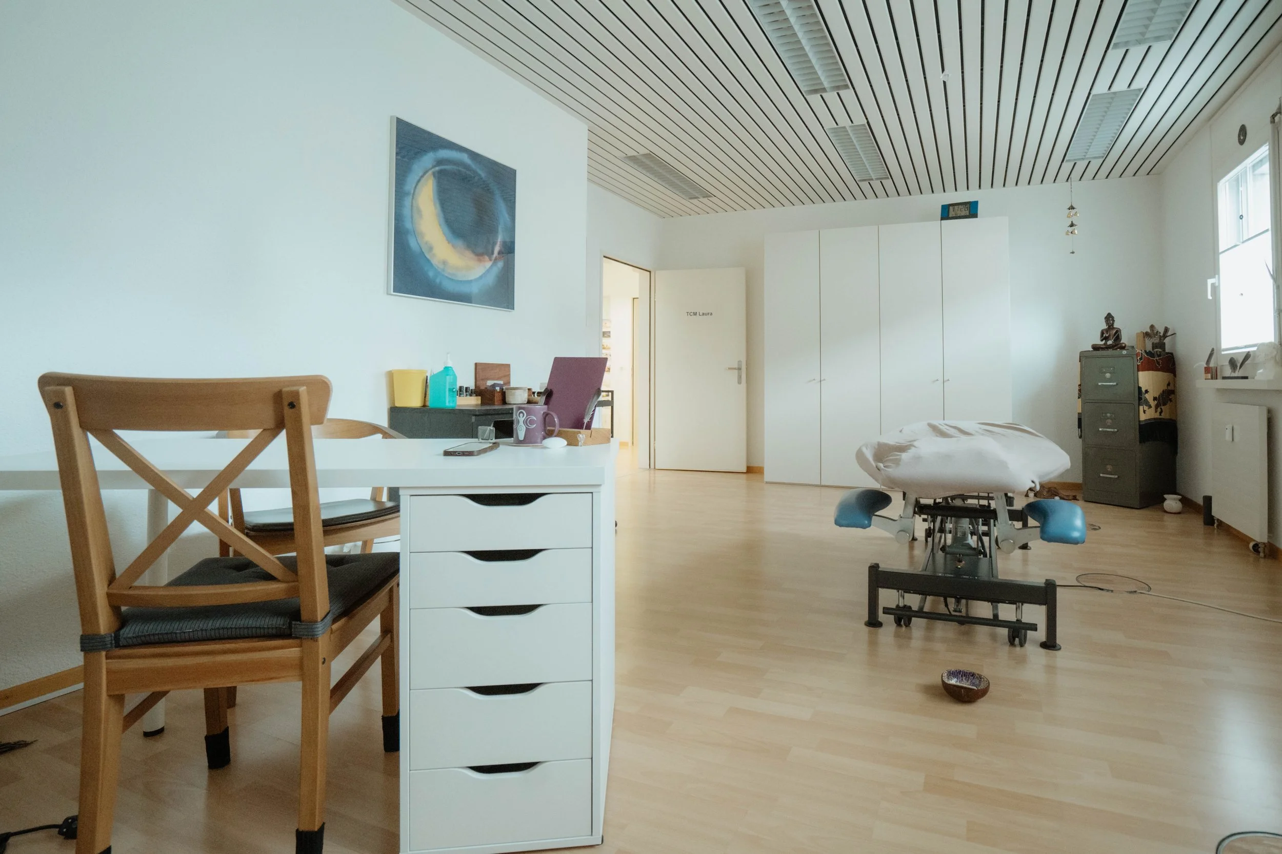 Ein Behandlungszimmer mit einem Behandlungstisch, einem Schreibtisch mit Stuhl, einem Regal mit dekorativen Gegenständen und einem Kunstbild an der Wand.