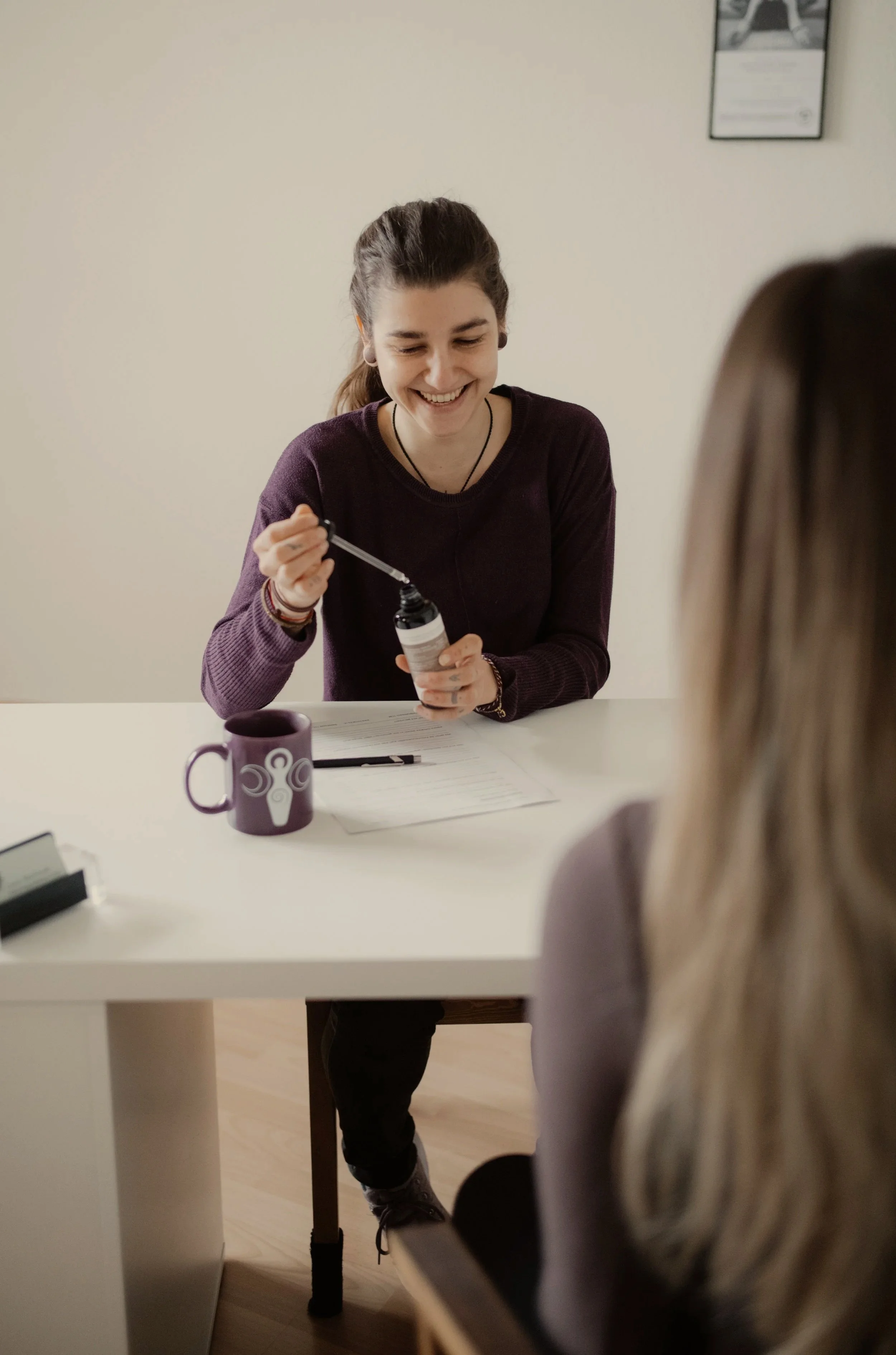 Eine lachende junge Frau, die an einem Behandlungstisch gegenüber einer anderen Person sitzt. Auf dem Tisch liegen Papier und ein Stift.