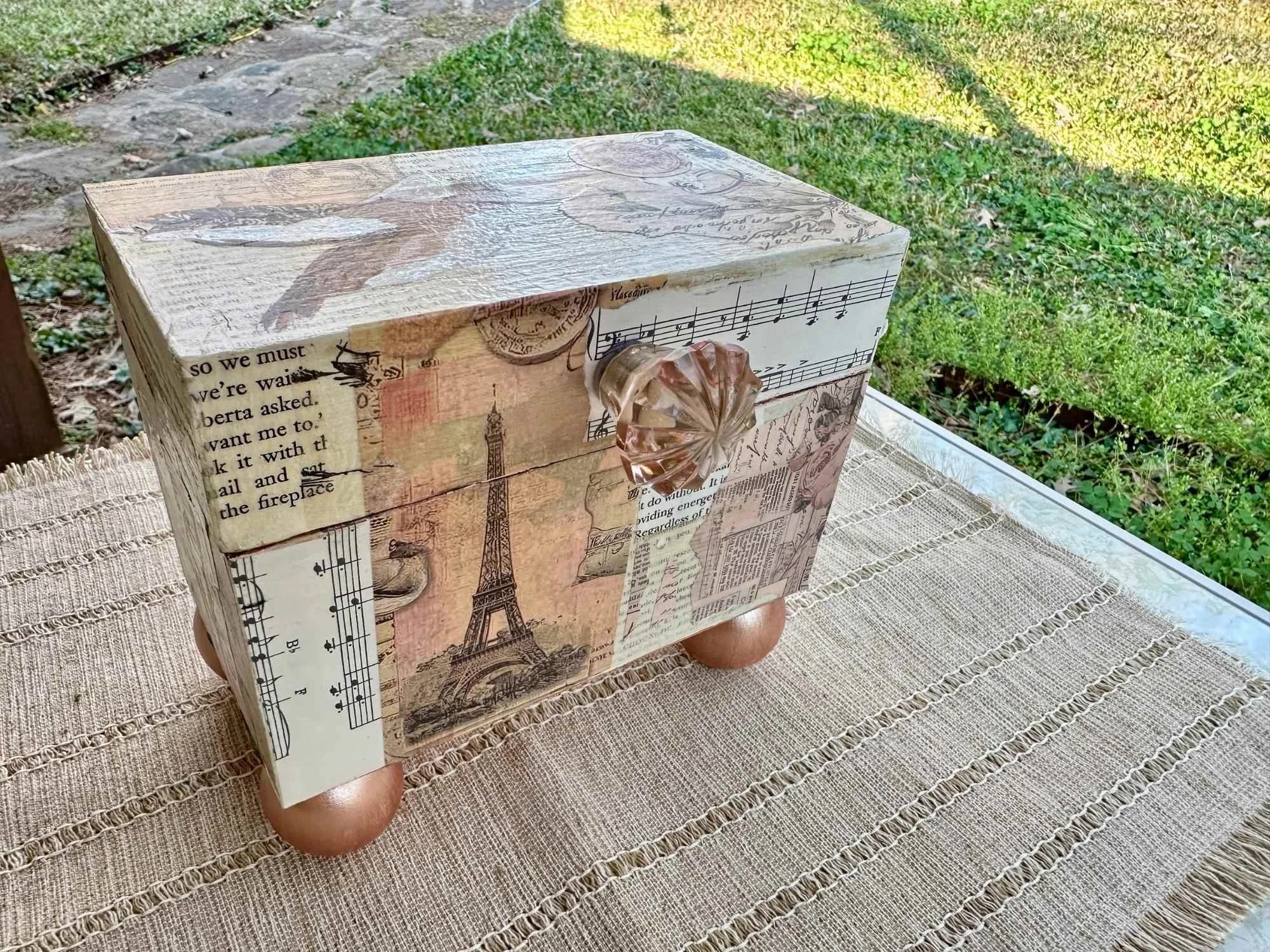 A decorative wooden box with vintage newspaper and sheet music prints, featuring Eiffel Tower illustrations and a crystal knob on the drawer, being held outdoors on a tablecloth with a grassy background.