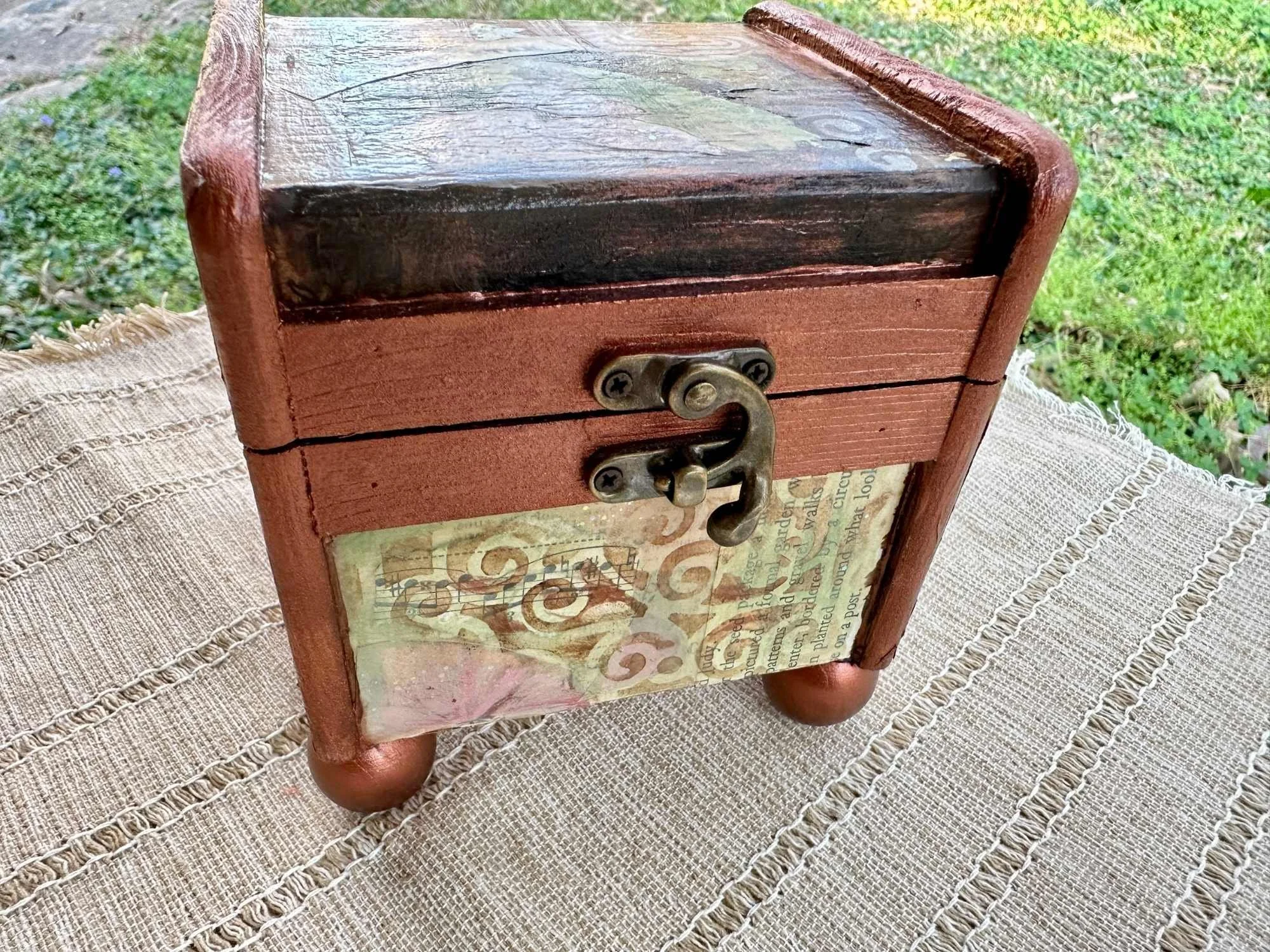 A small wooden box with a rusty latch on the front, sitting on a fabric tablecloth. The box has a vintage poster or paper with decorative text and design on one side.