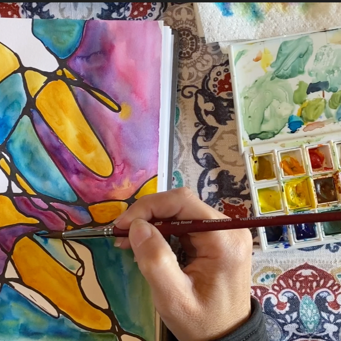 Person painting colorful stained glass-style butterfly design with a watercolor set on a table.