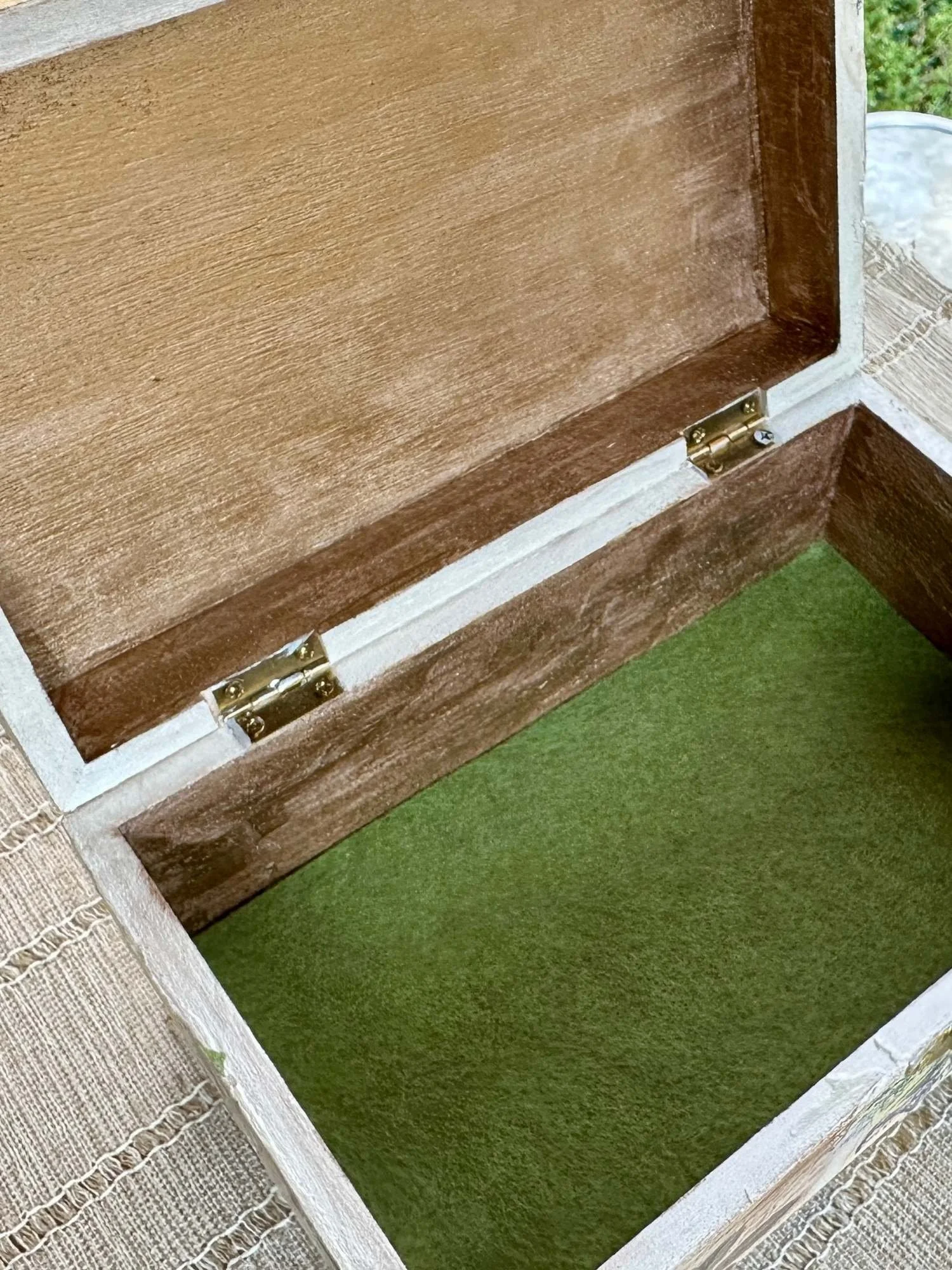 Close-up of an open wooden box with brass hinges, resting on a green surface next to a woven fabric and stone wall.