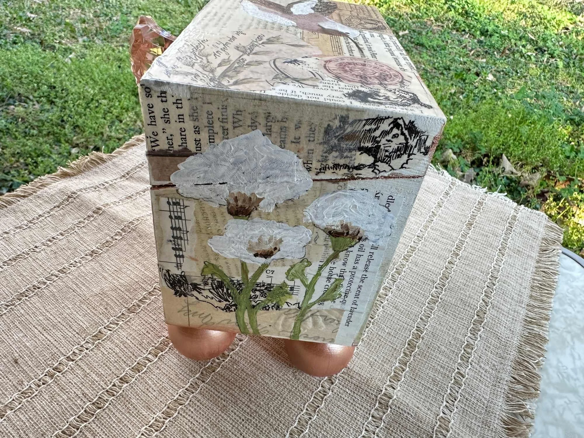 A decorated book with vintage and botanical illustrations, text, and musical notes, held in a person's hand outdoors, placed on a beige woven cloth. The background shows green grass and foliage.