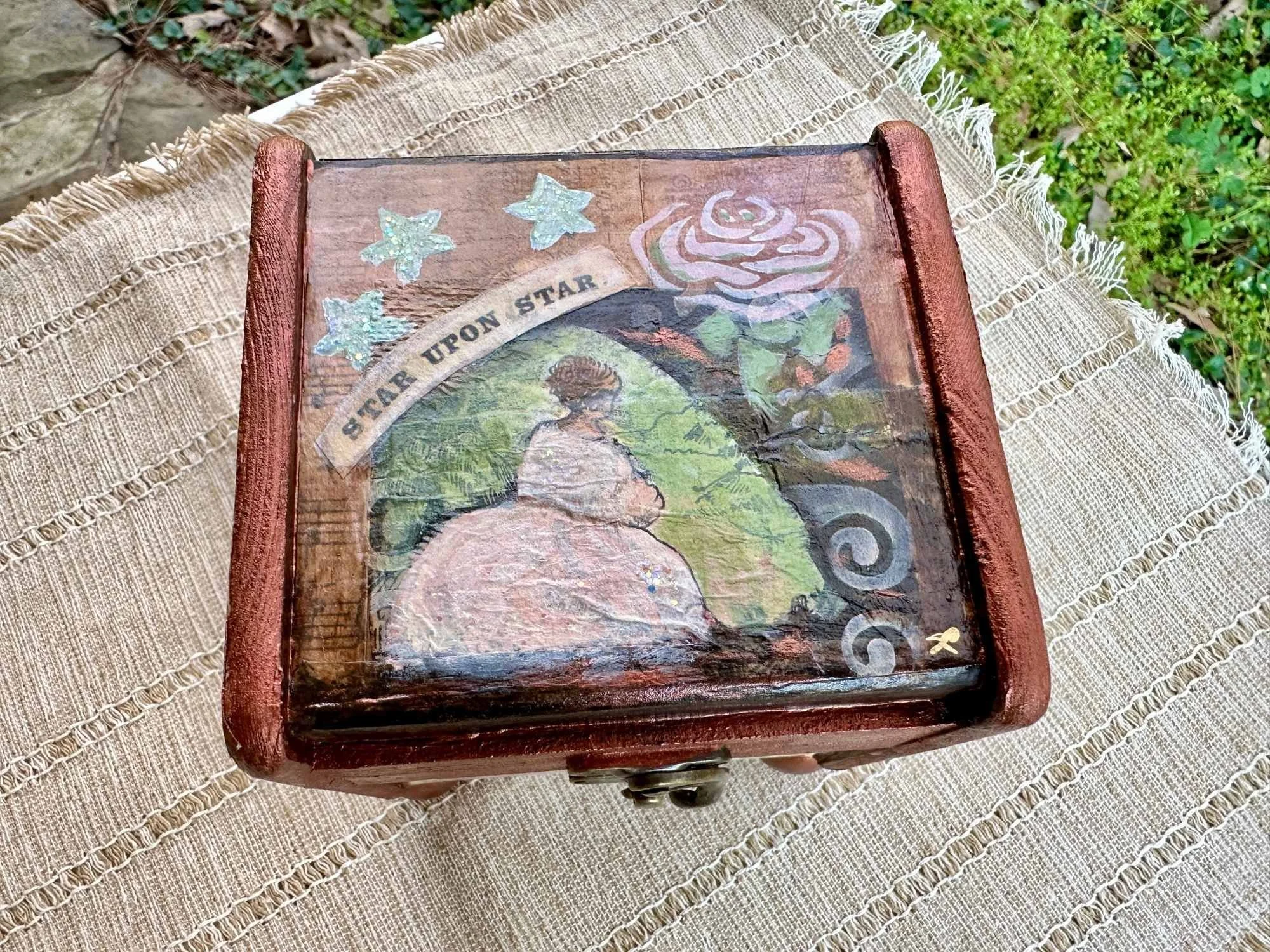 A wooden box with artwork, stickers, and a sticker quote reading 'Start your own star.' The box has a red-brown finish and is placed on a cream-colored woven cloth outdoors on grass and rocks.