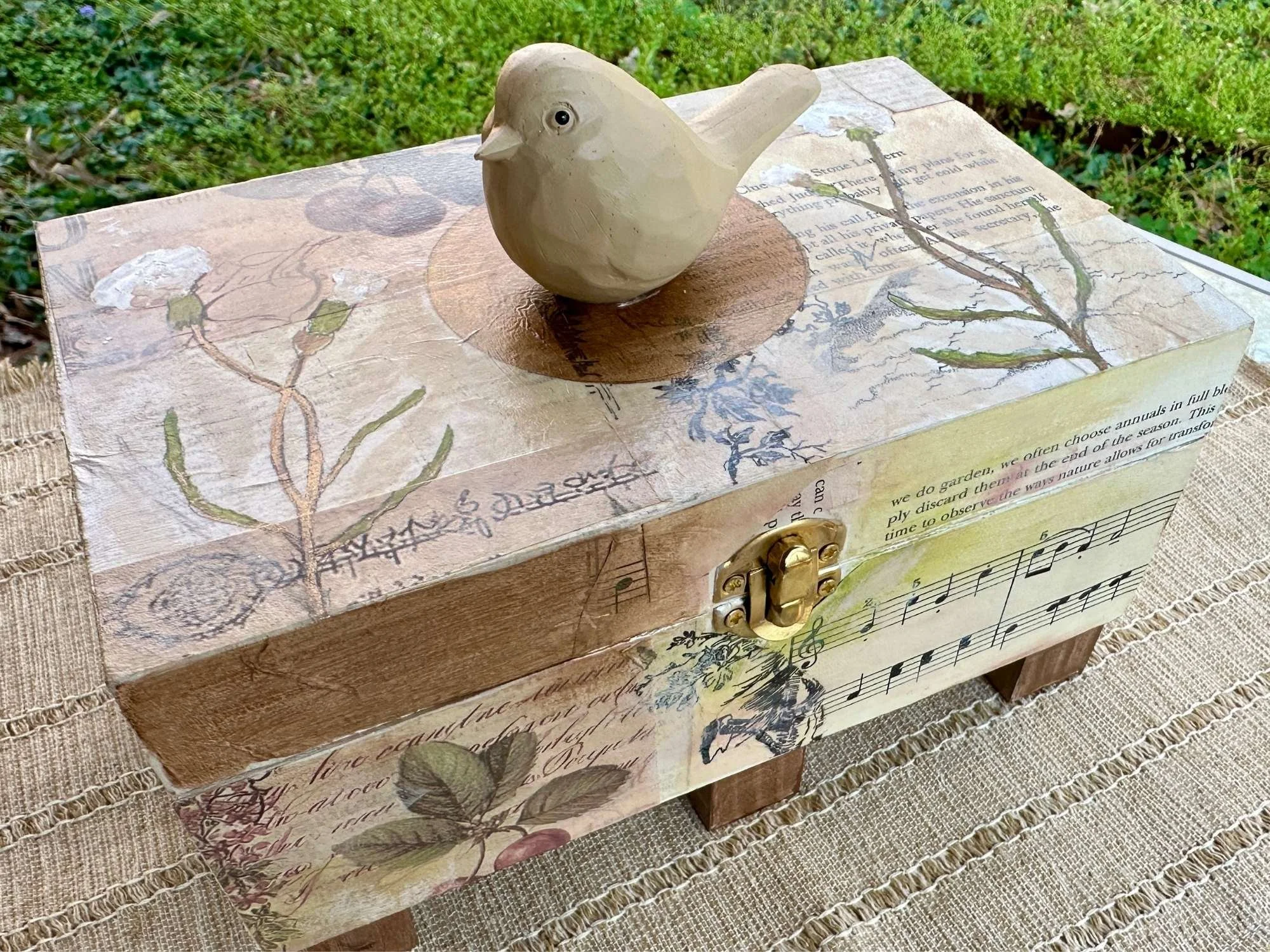 Decorative wooden jewelry box with a carved bird on top, featuring a cream-colored finish and musical sheet design on its surface, placed on a textured fabric surface.