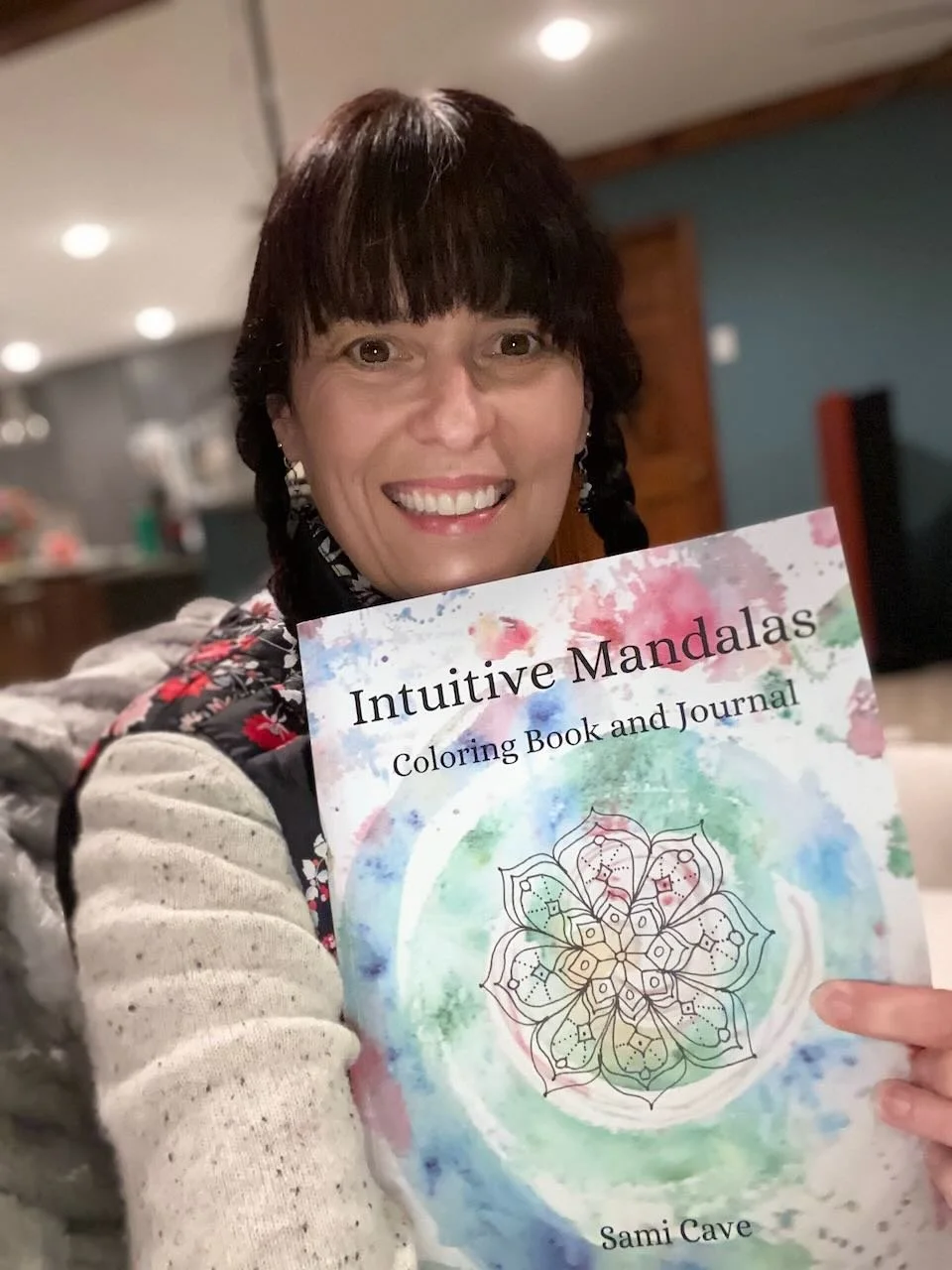 A woman with dark hair and bangs smiling while holding a colorful book titled "Intuitive Mandalas Coloring Book and Journal" by Sami Cave, indoors with blurred background.