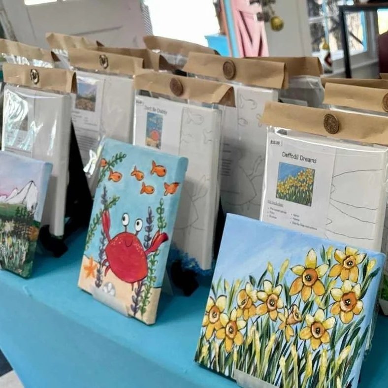 Paintings of nature scenes and a cartoon crab on display at an art fair or market.