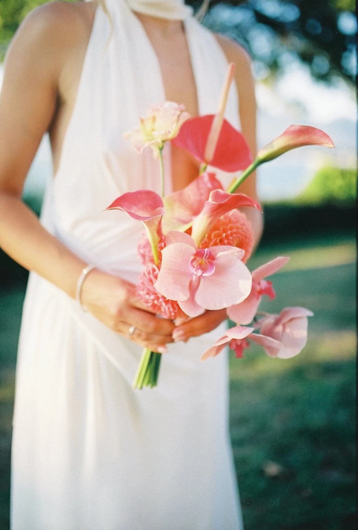 A woman in a white dress holds a bouquet of pink flowers with green stems, including orchids and other tropical blooms, outdoors with a blurred natural background.