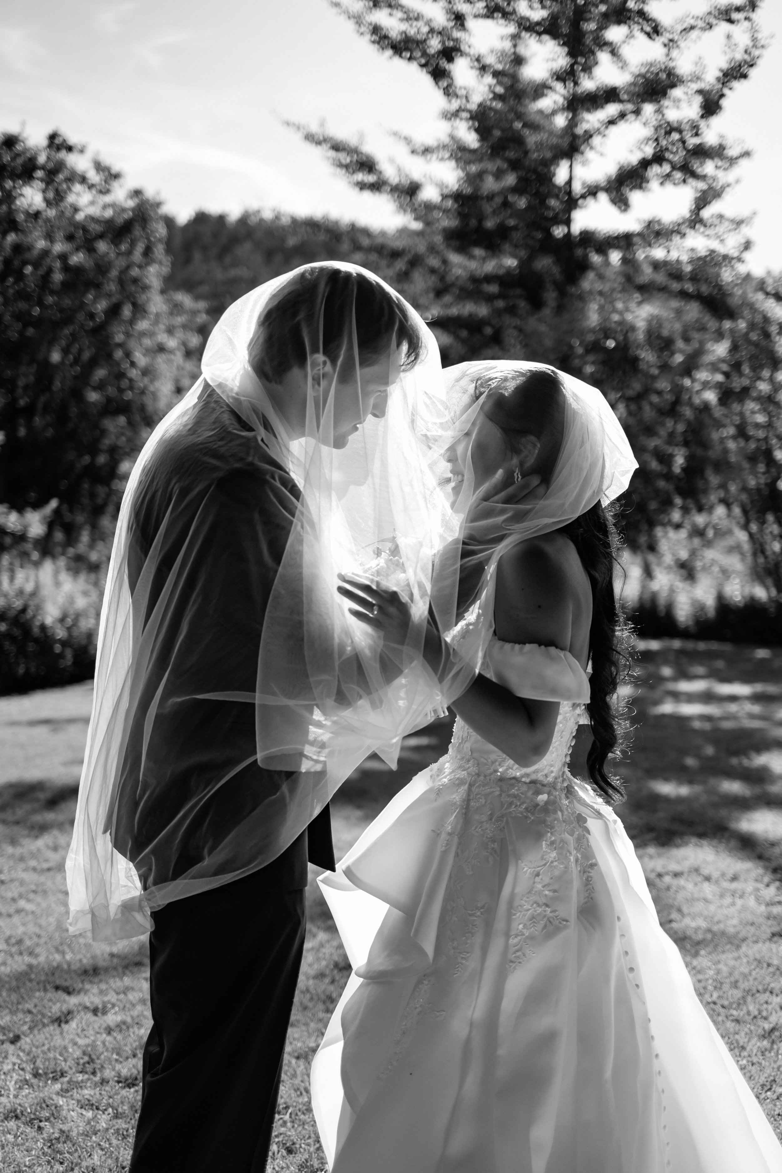 Black and white photo of a bride and groom under a wedding veil outside, smiling and close together, with trees and sunlight in the background.