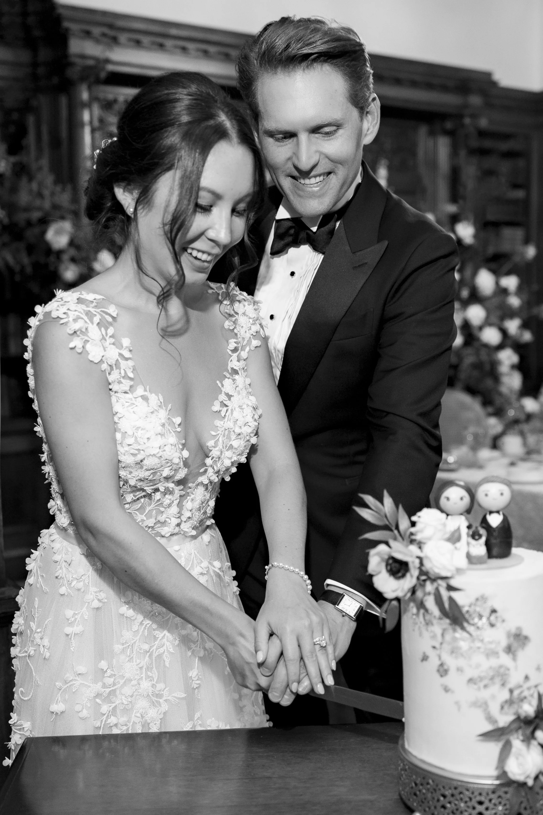 A bride and groom in wedding attire cutting a wedding cake together, smiling, with a decorated table in the background.