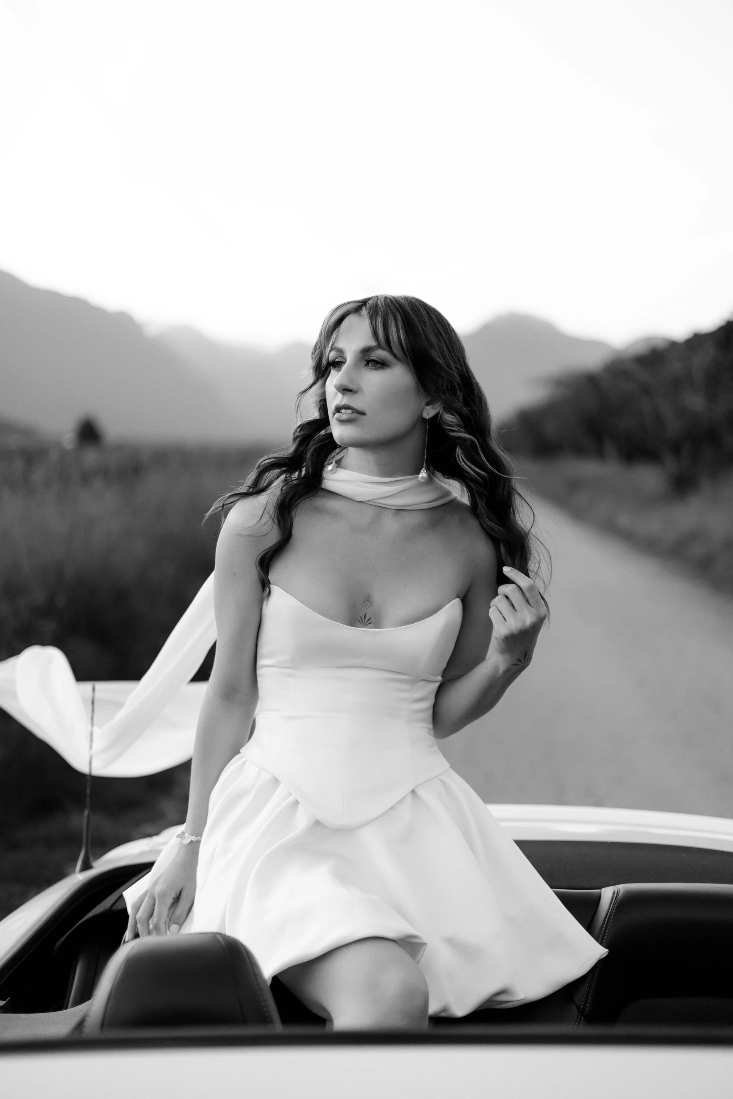 A woman in a white strapless dress with a scarf around her neck, sitting on the back seat of a convertible car in a rural setting with mountains in the background.