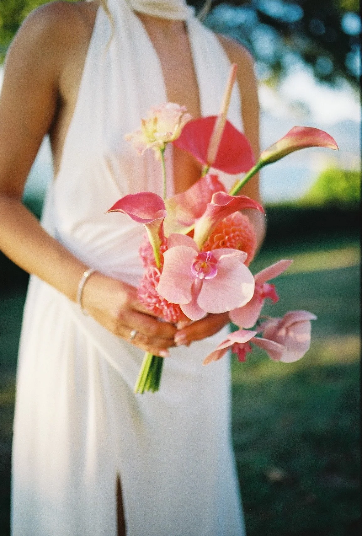 A woman in a white dress holding a bouquet of pink and peach flowers with green leaves in an outdoor setting with blurred greenery in the background.