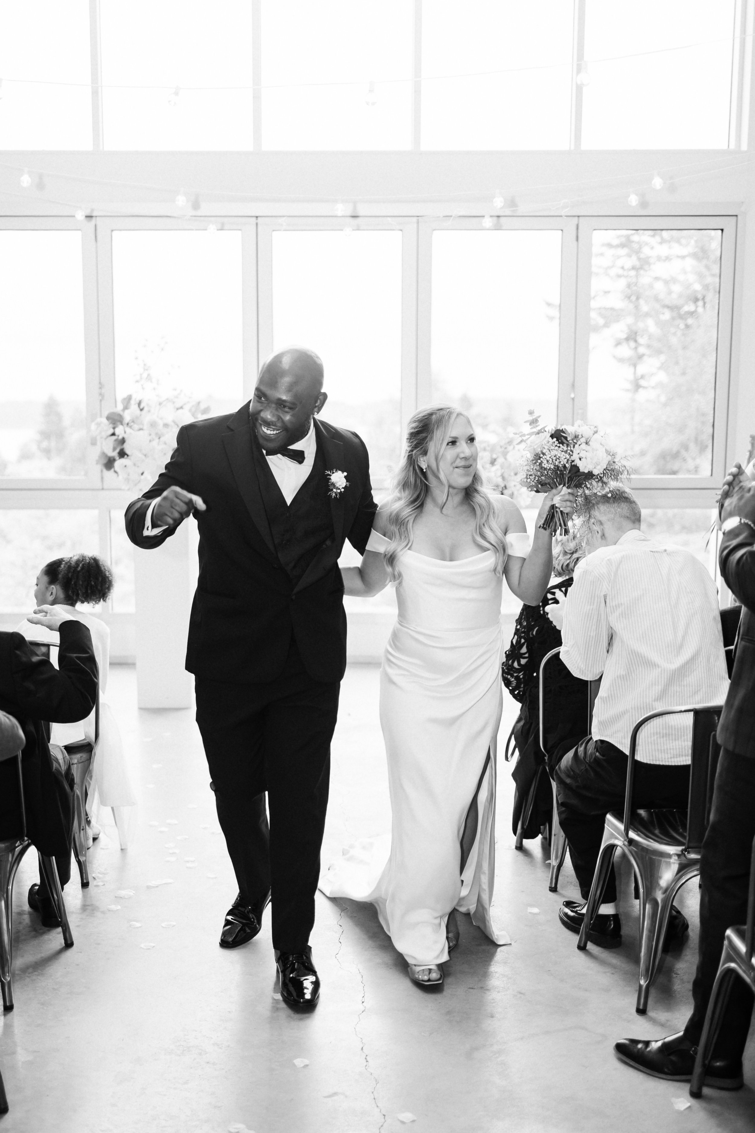 A happy wedding couple walking through a reception hall, with guests seated on either side, in a black and white photo. The man is in a tuxedo, and the woman is in a white wedding dress holding a bouquet.