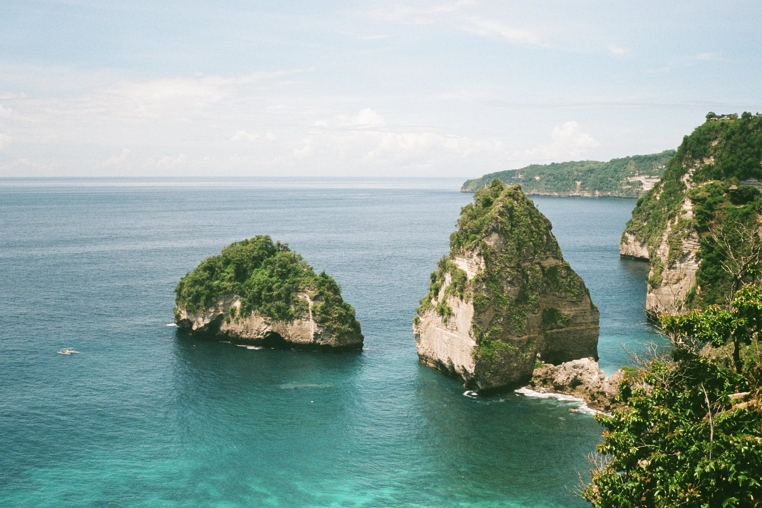 Three large rocky islands covered in greenery emerging from the ocean, with a cloudy sky above and cliffs in the background.