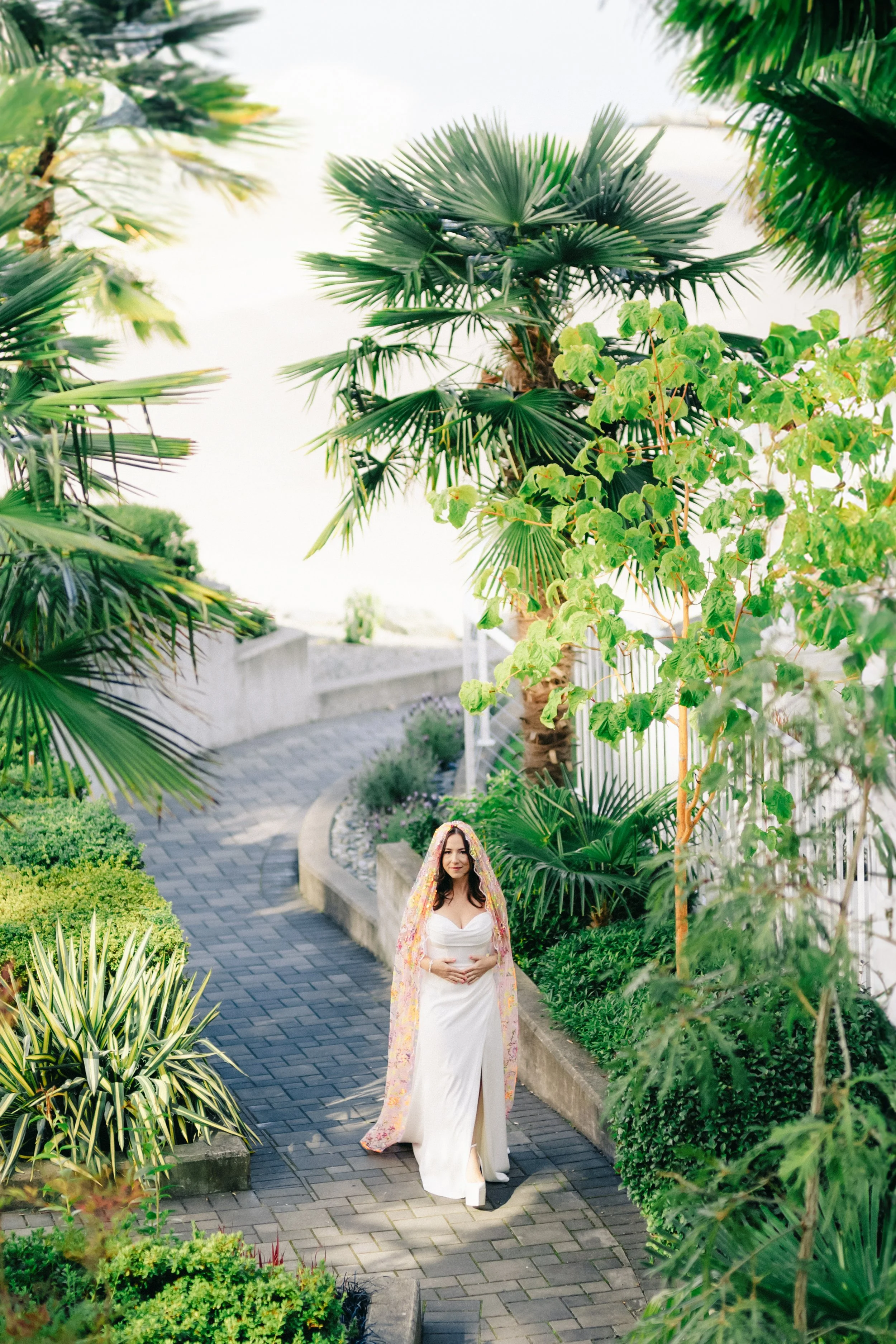 A woman dressed in white walking along a garden path with lush green tropical plants and trees surrounding her.