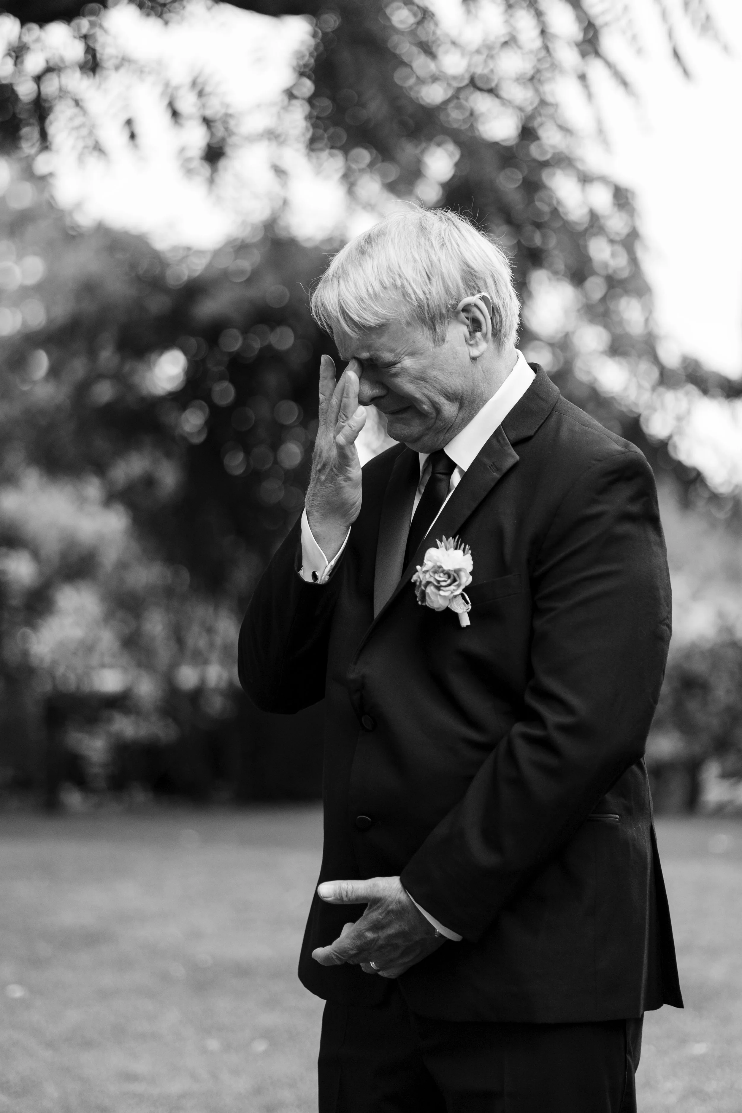 An elderly man in a suit with a boutonniere is visibly emotional, with his hand on his face and eyes closed, standing outdoors in a grassy area with blurred trees in the background.