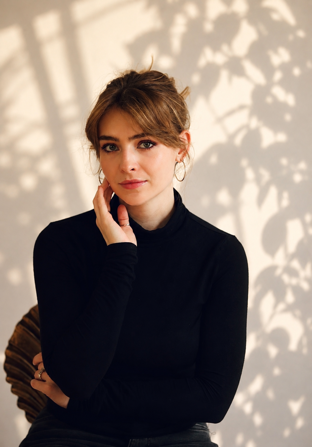A woman with light brown hair and blue eyes wearing a black turtleneck, posing with her hand near her face, in front of a background with shadowed leaf patterns.