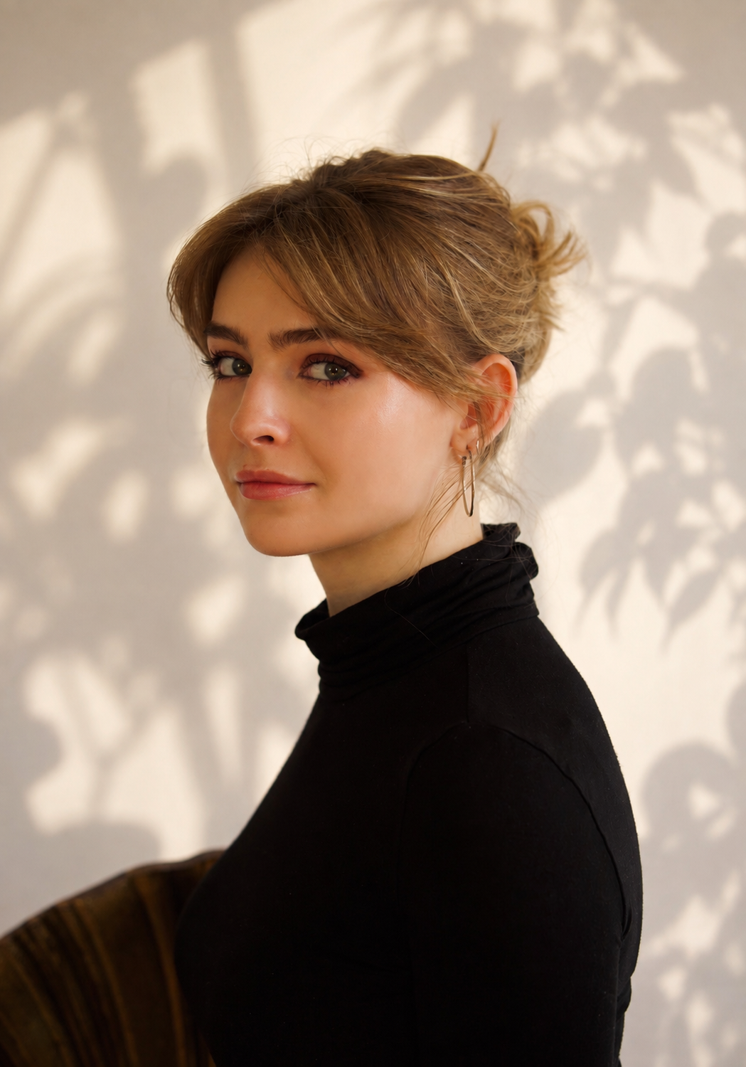 A woman with short, wavy brown hair and light skin, wearing a black turtleneck sweater and hoop earrings, looking slightly over her shoulder against a background with shadowed leaf patterns.
