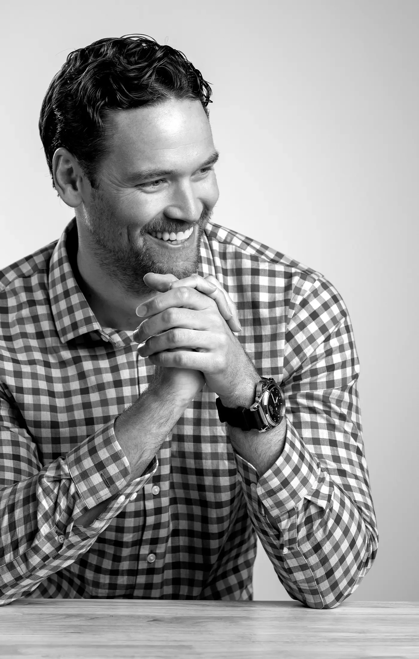 Black and white photo of Kyle with dark, wavy hair and beard, smiling, wearing a checkered shirt and a watch, hands clasped, sitting at a table against a plain background.
