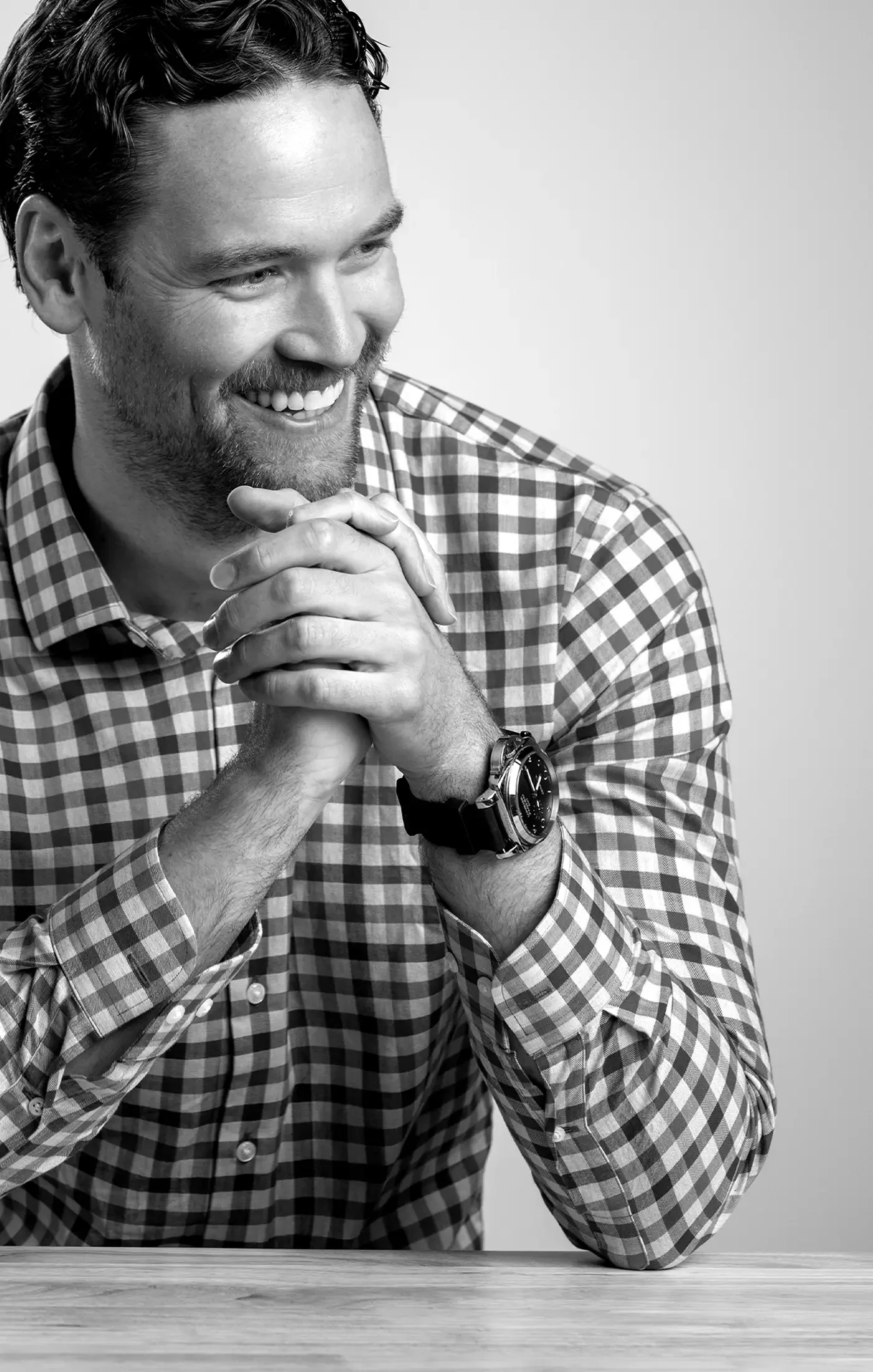 Black and white photo of Kyle Casteel smiling, sitting at a table with his hands clasped together, wearing a checkered shirt and a watch.