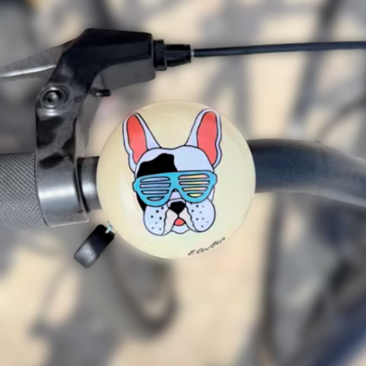 Close up of bike bell attached to handle bars of a bike. Bike bell is round and has an animation of a french bulldog wearing blue avaiator sunglasses on a light cream background