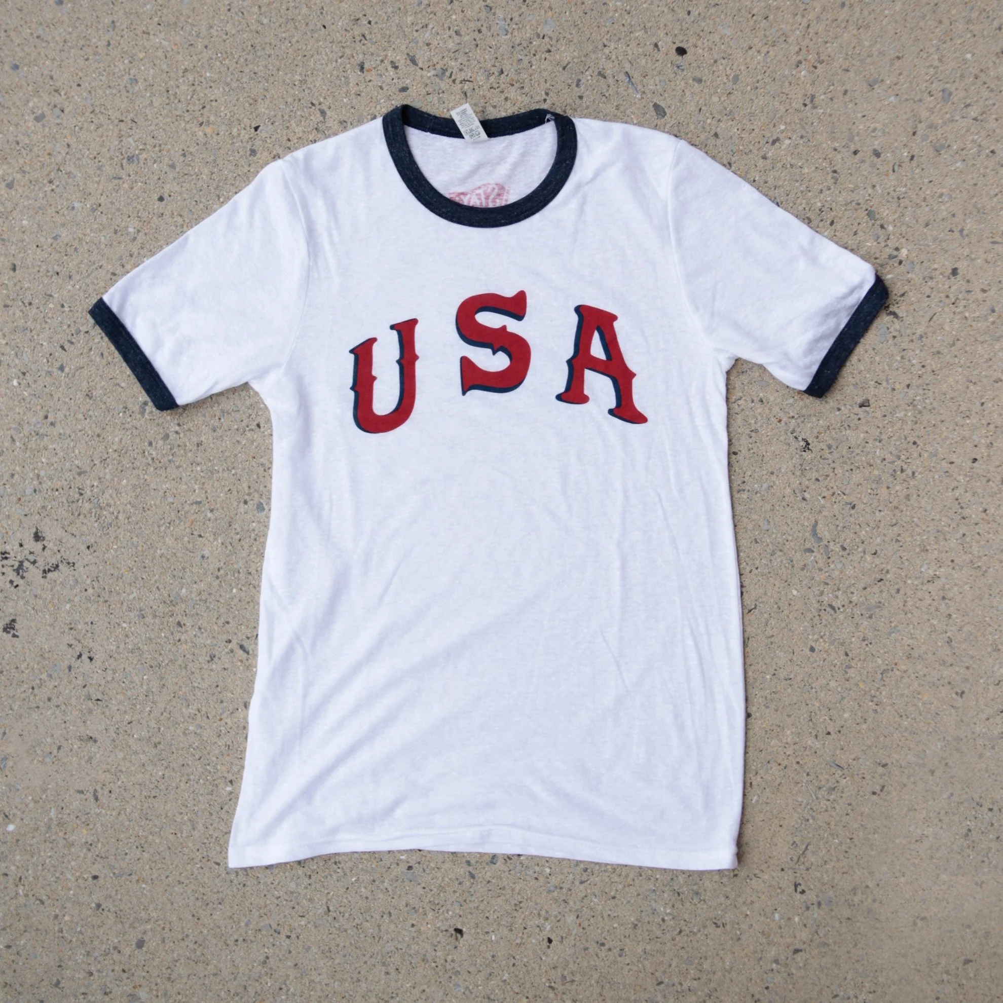 White ringer tee with blue accents and USA printed in a retro font of red ink and a blue shadow.