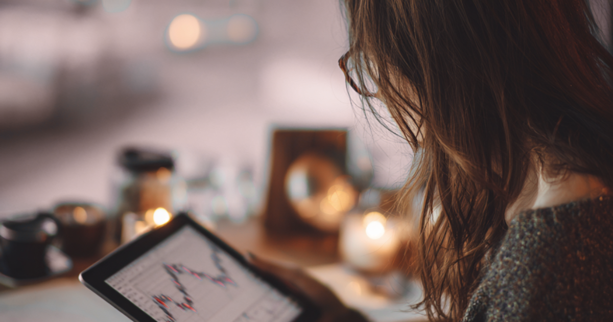 A woman looking at a tablet with financial graphs on the screen, with candles and blurred objects in the background.