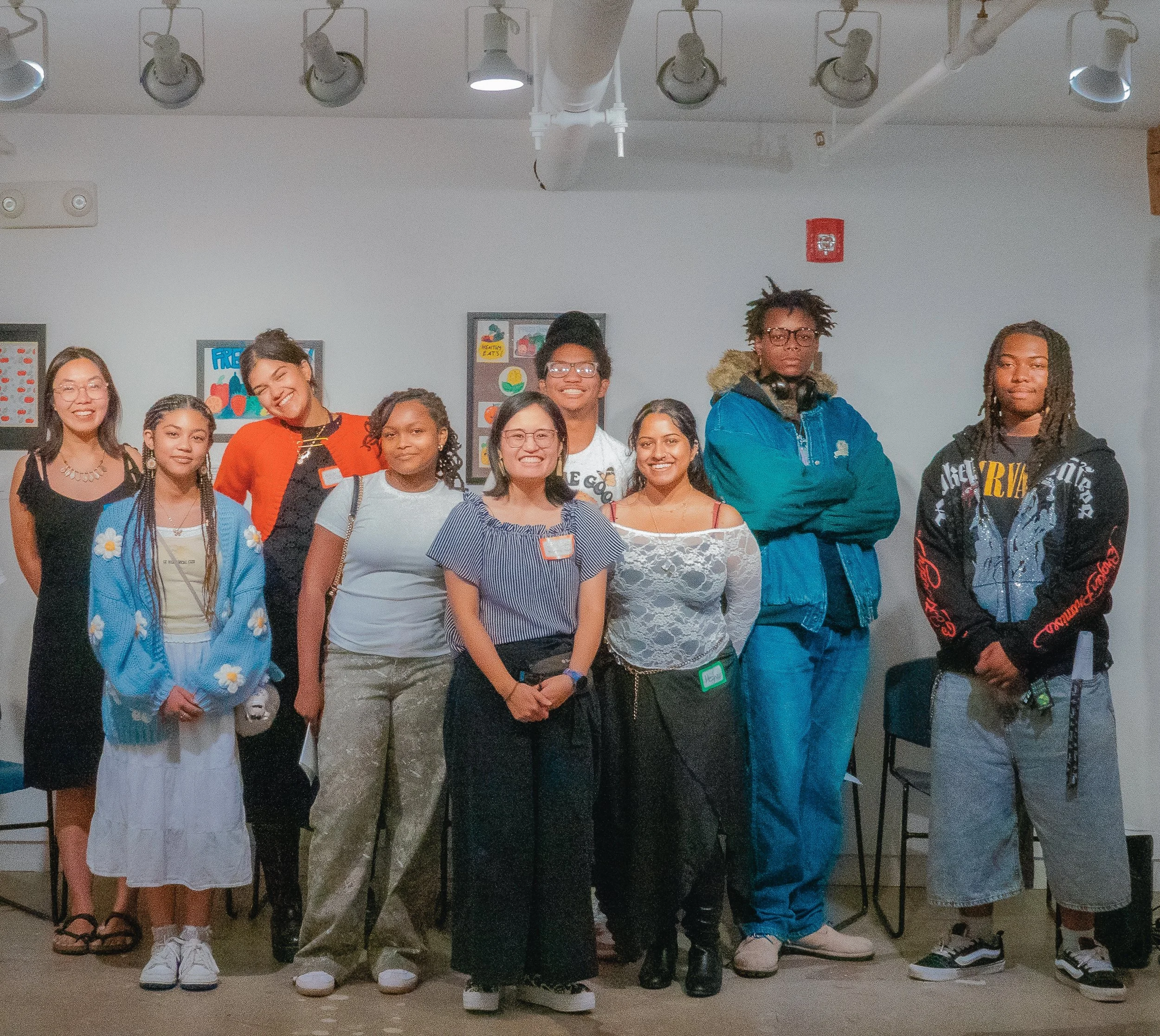 Group of ten young adults standing indoors, smiling for a photo. The background features framed artwork and a white wall with ceiling lights.