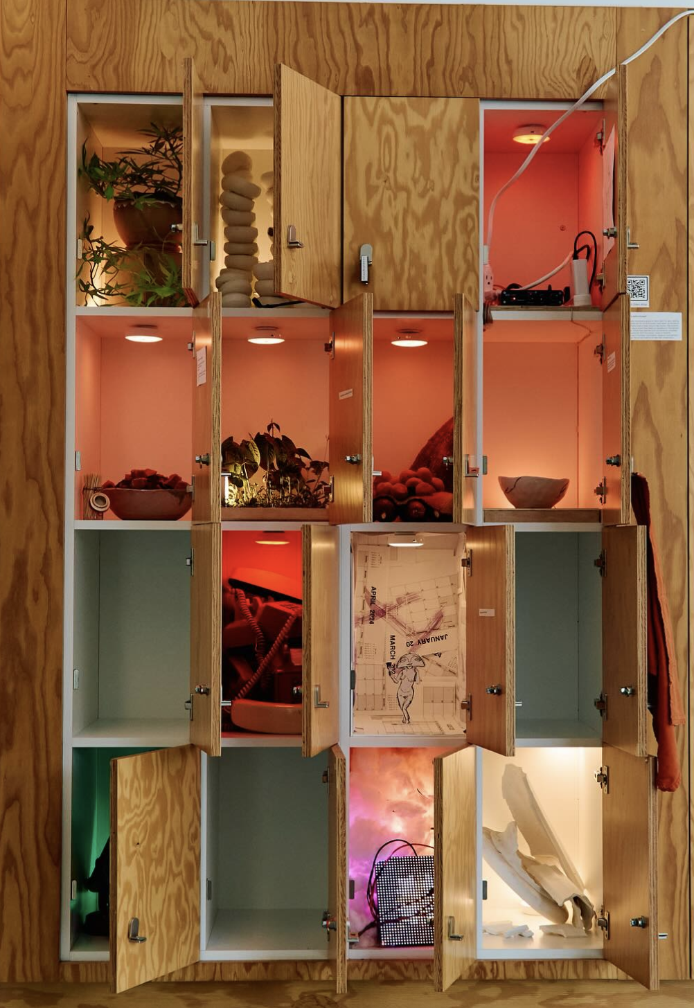 A wooden wall with multiple open shelves, each containing different objects including plants, decorative stones, a kaleidoscope, a framed illustration, and various electronic devices with colorful lighting.