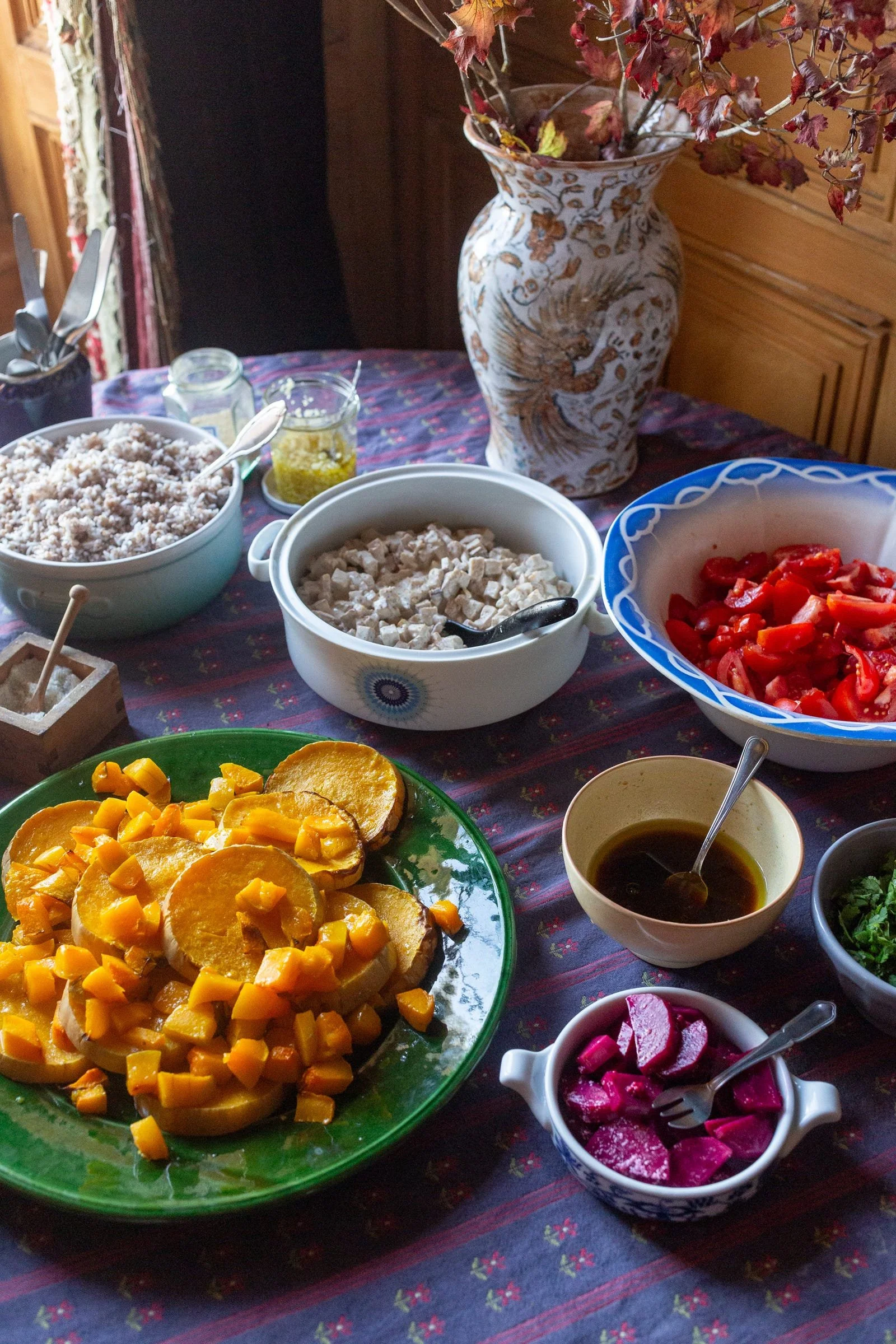 A table set with various bowls of food including diced vegetables, a salad, and sauces, with a vase of dried flowers in the background.
