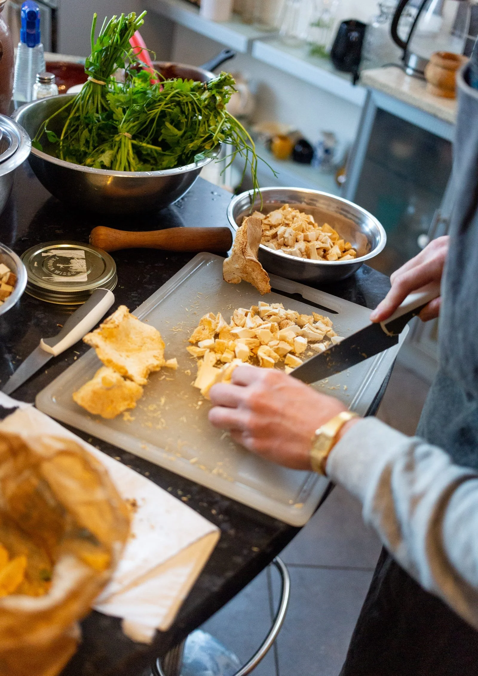 Person chopping cooked chicken on a cutting board in a kitchen, with fresh herbs and kitchen utensils around.