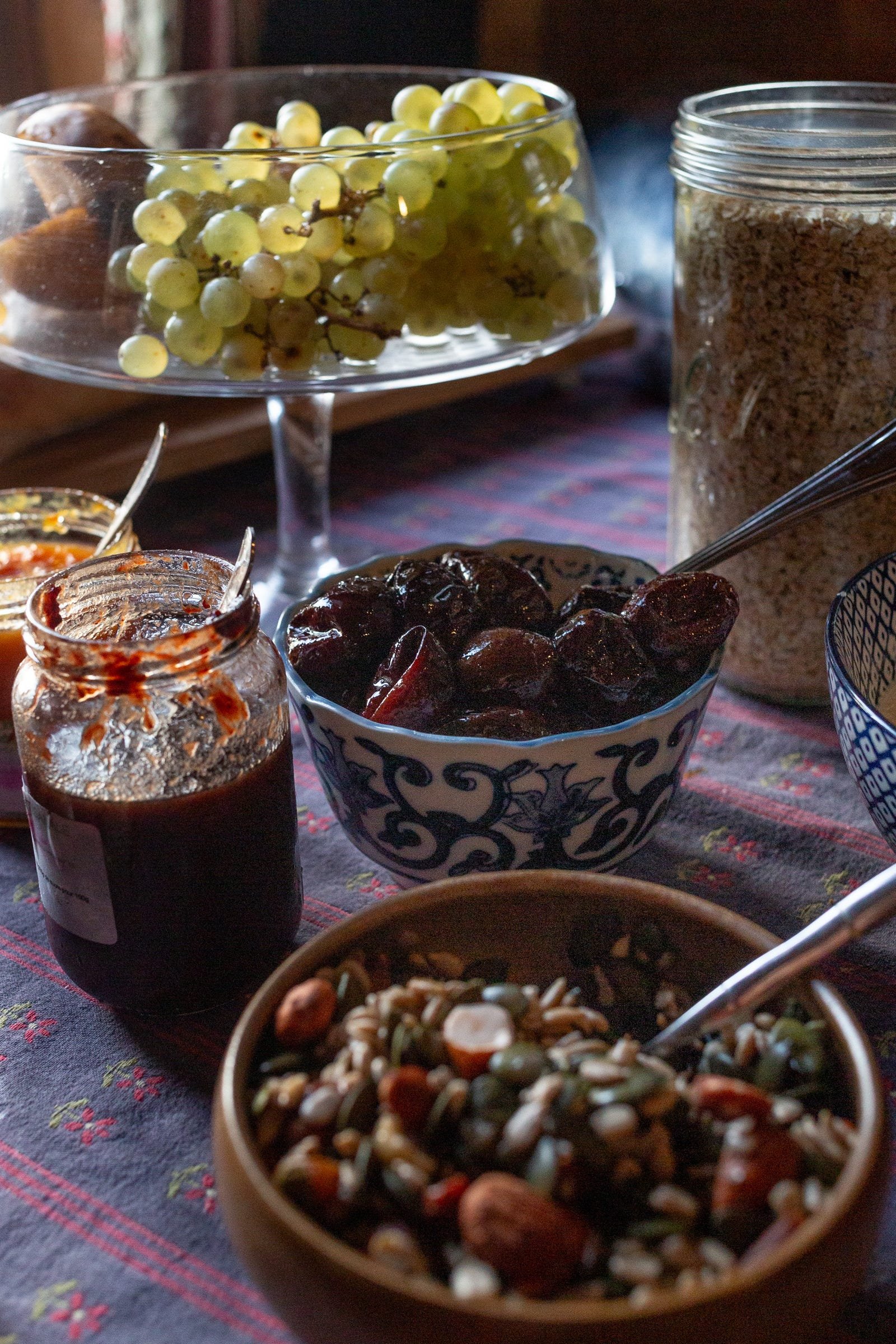 A table with various bowls and jars of food, including a bowl of mixed nuts, a bowl of dried dates, a jar of jam, a jar of cereal, a bowl of green grapes, and a jar of syrup.