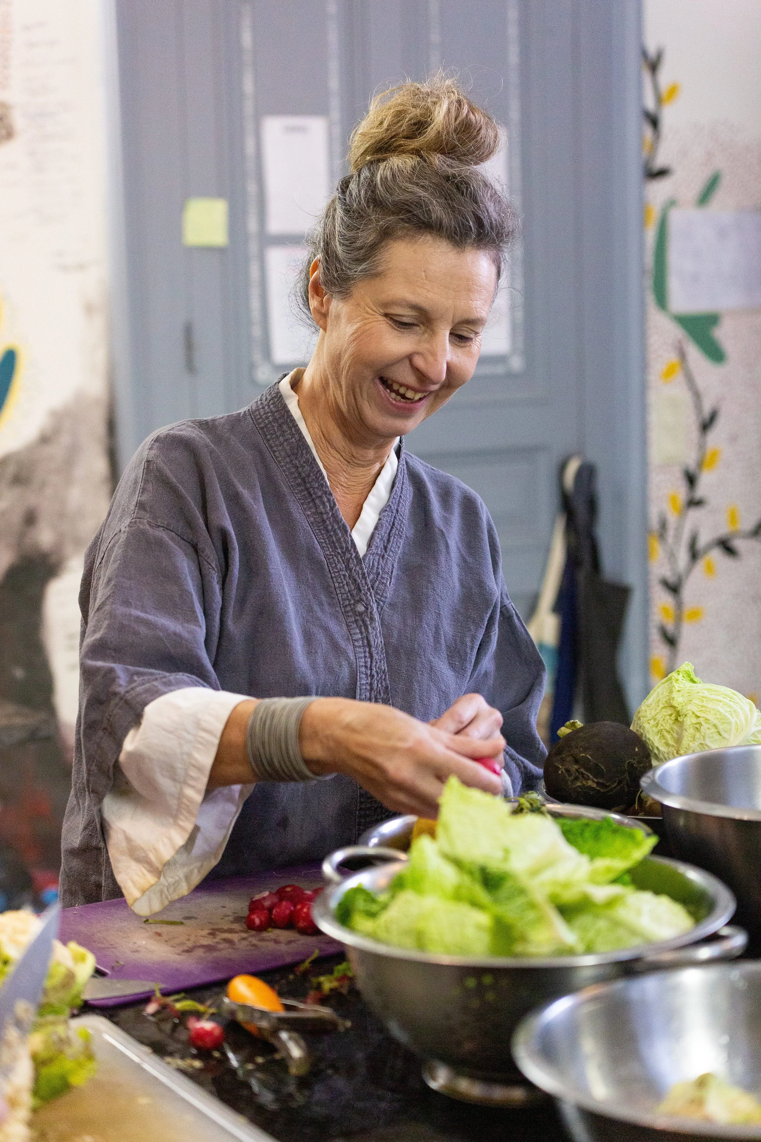 A woman with gray hair tied in a bun, wearing a gray apron, preparing vegetables in a kitchen.