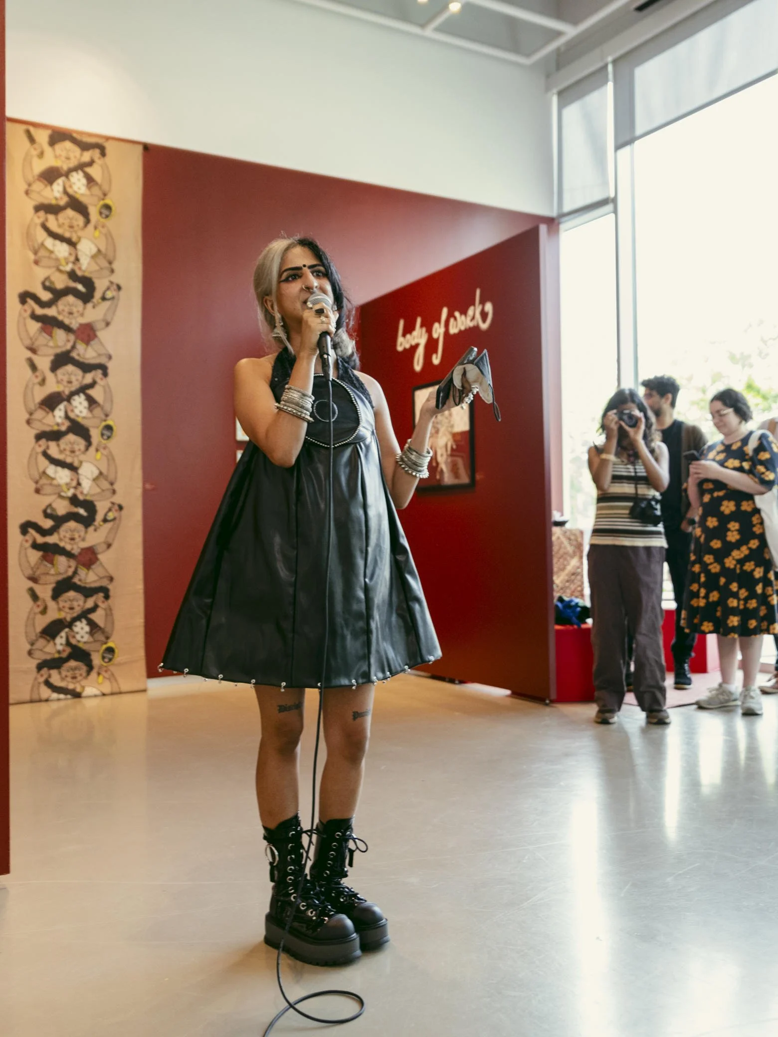 A young woman with dyed hair and tattoos, dressed in a black dress and platforms, speaking into a microphone at an indoor event with a red wall and crowd in the background.
