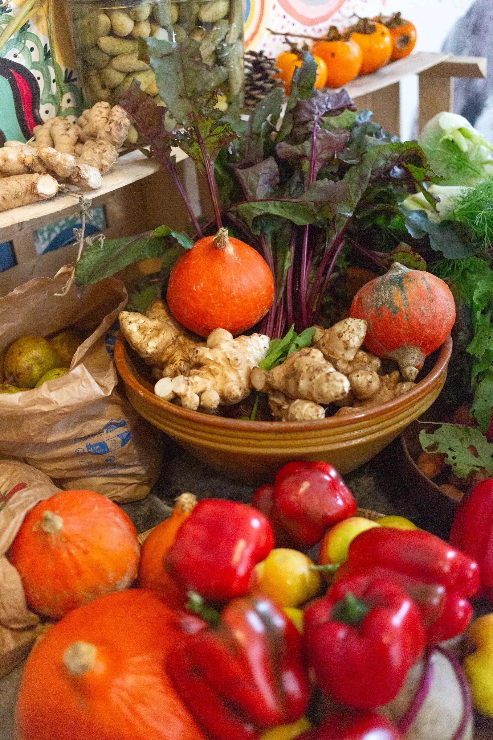 Display of fresh vegetables including pumpkins, ginger roots, red and yellow apples, and red bell peppers on a table at a farmers market.