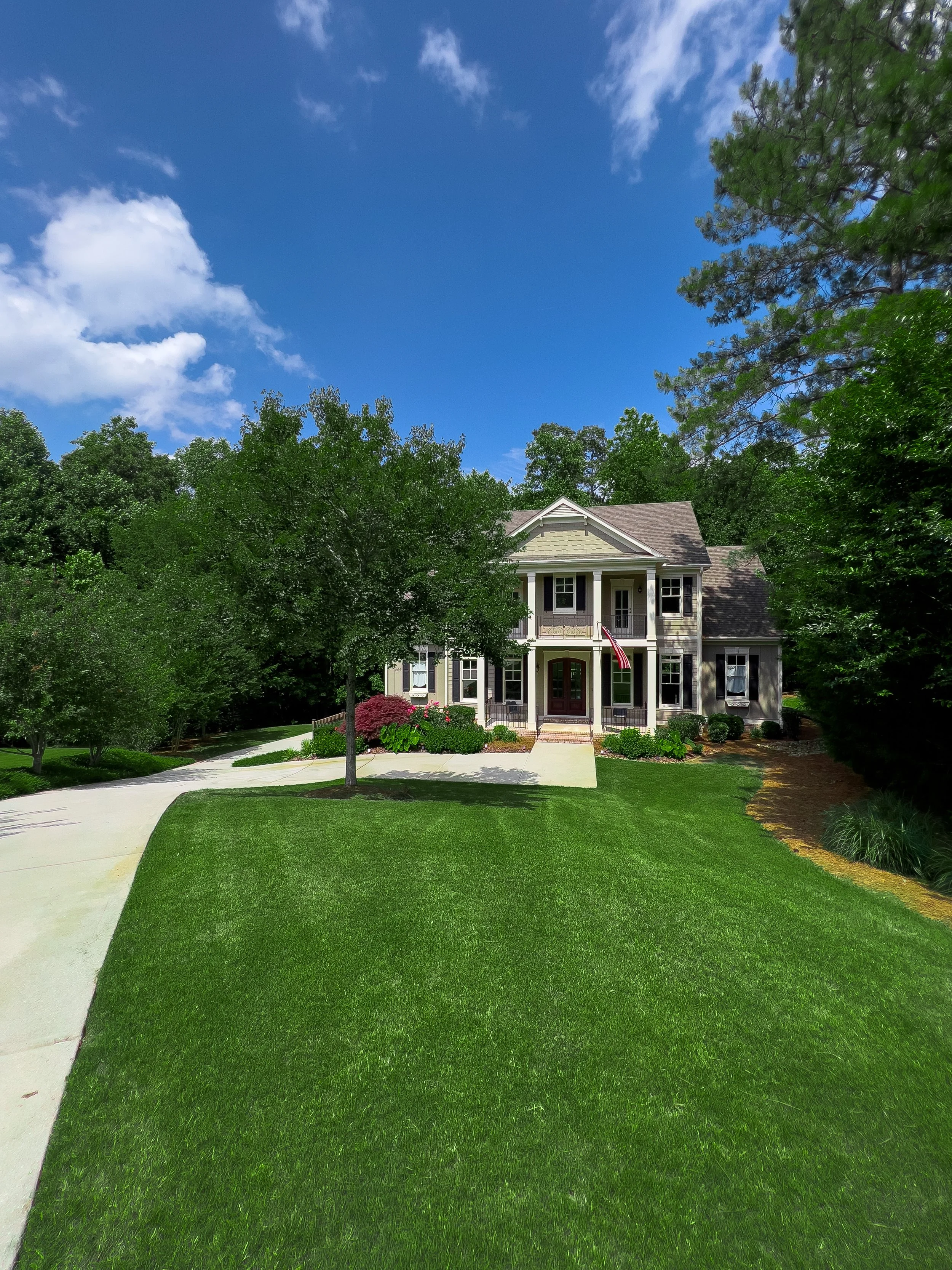 Front view of a large, two-story house with a well-maintained lawn, trees, and a driveway on a sunny day with blue sky and scattered clouds.