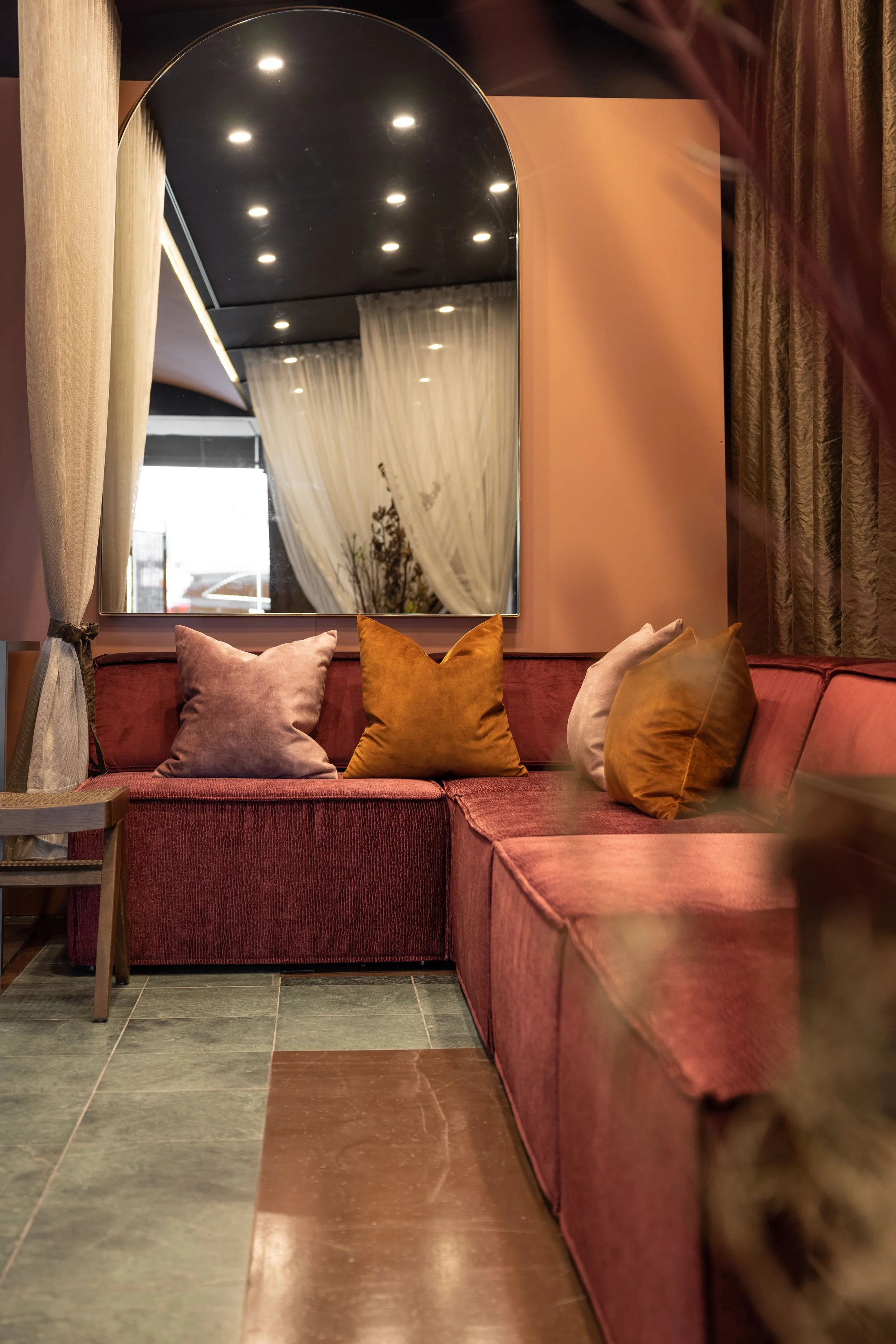 A cozy seating area with a red velvet sofa decorated with various pink, orange, and beige pillows. Behind the sofa is a large mirror reflecting ceiling lights, a window with curtains, and a decorated wall. A small wooden side table is near the sofa.